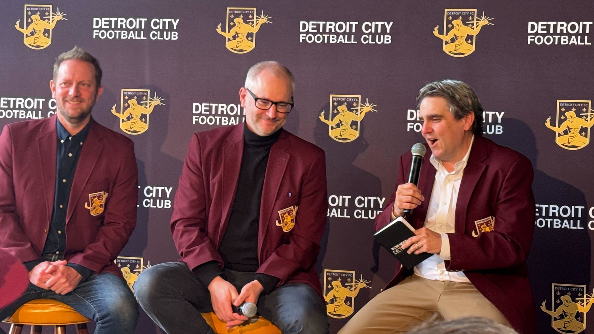 Three Detroit City FC executives in maroon blazers talk against a backdrop of DCFC's logo.