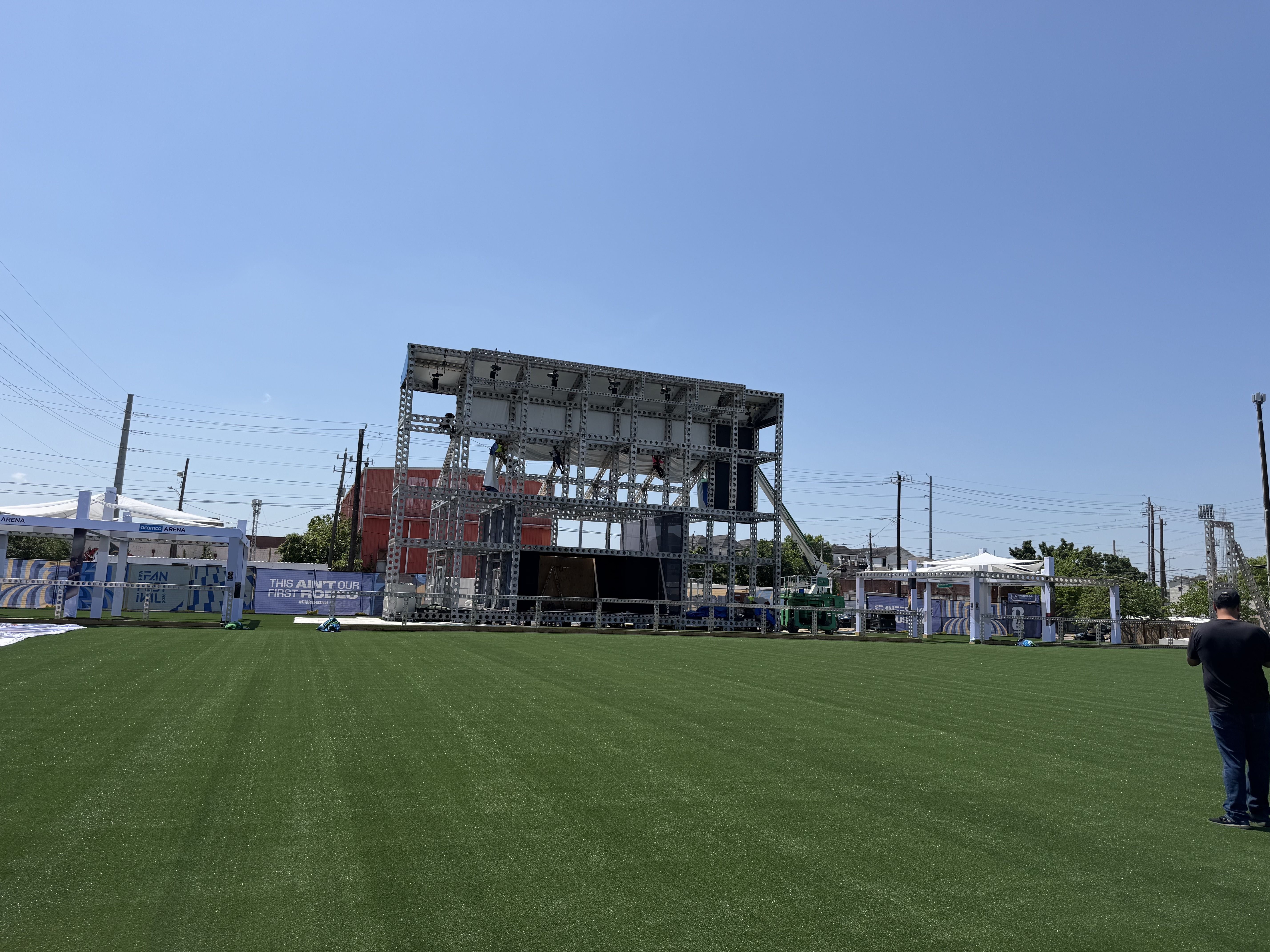 Bright blue sky over a large metal scaffolding structure on a green artificial turf field. White tents and banners line the foreground, with a person in dark clothing near the edge.