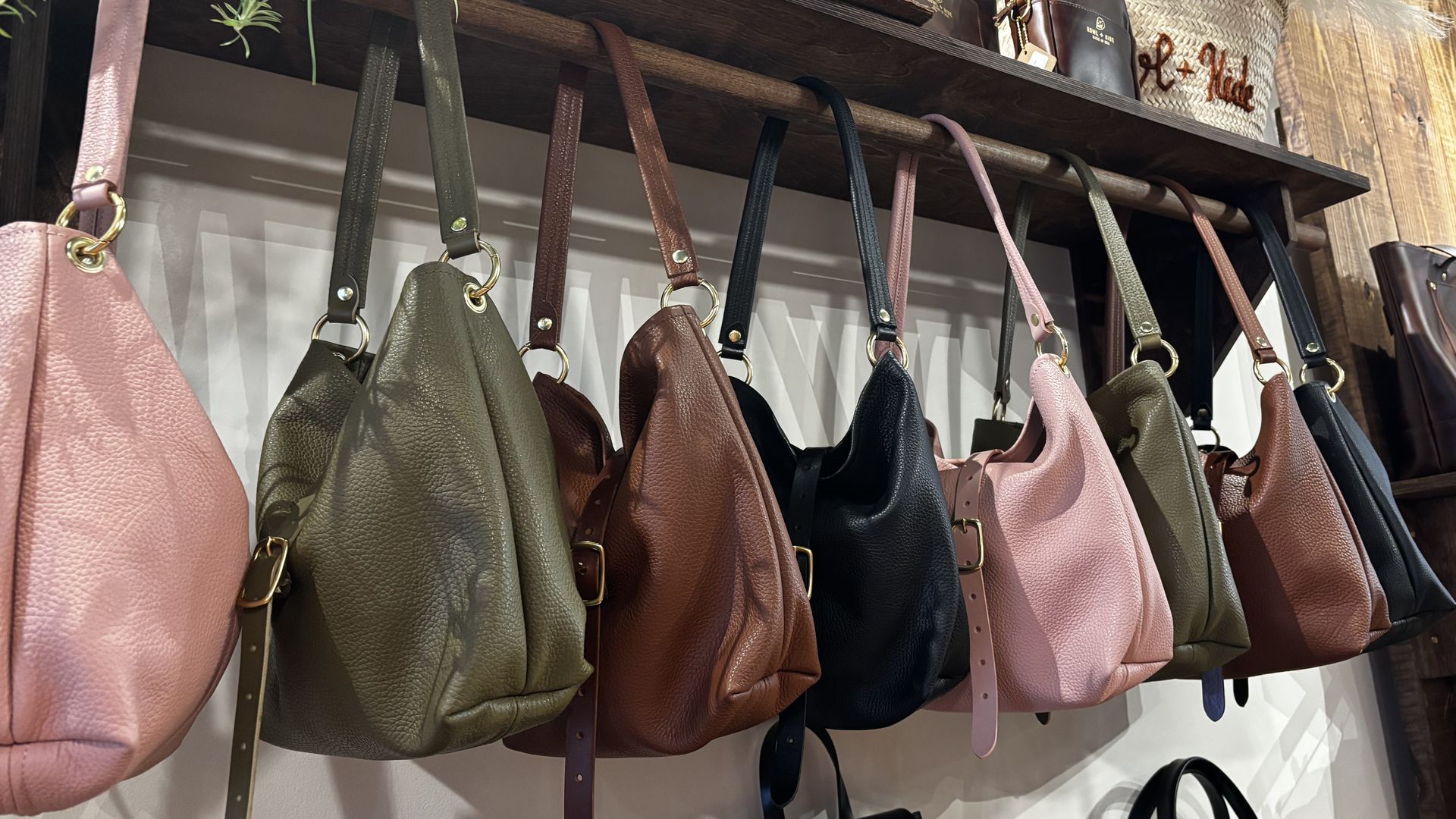 Shelf with hanging leather handbags in pink, olive green, brown, and black, alongside a woven basket with pampas grass inside a wooden interior store.