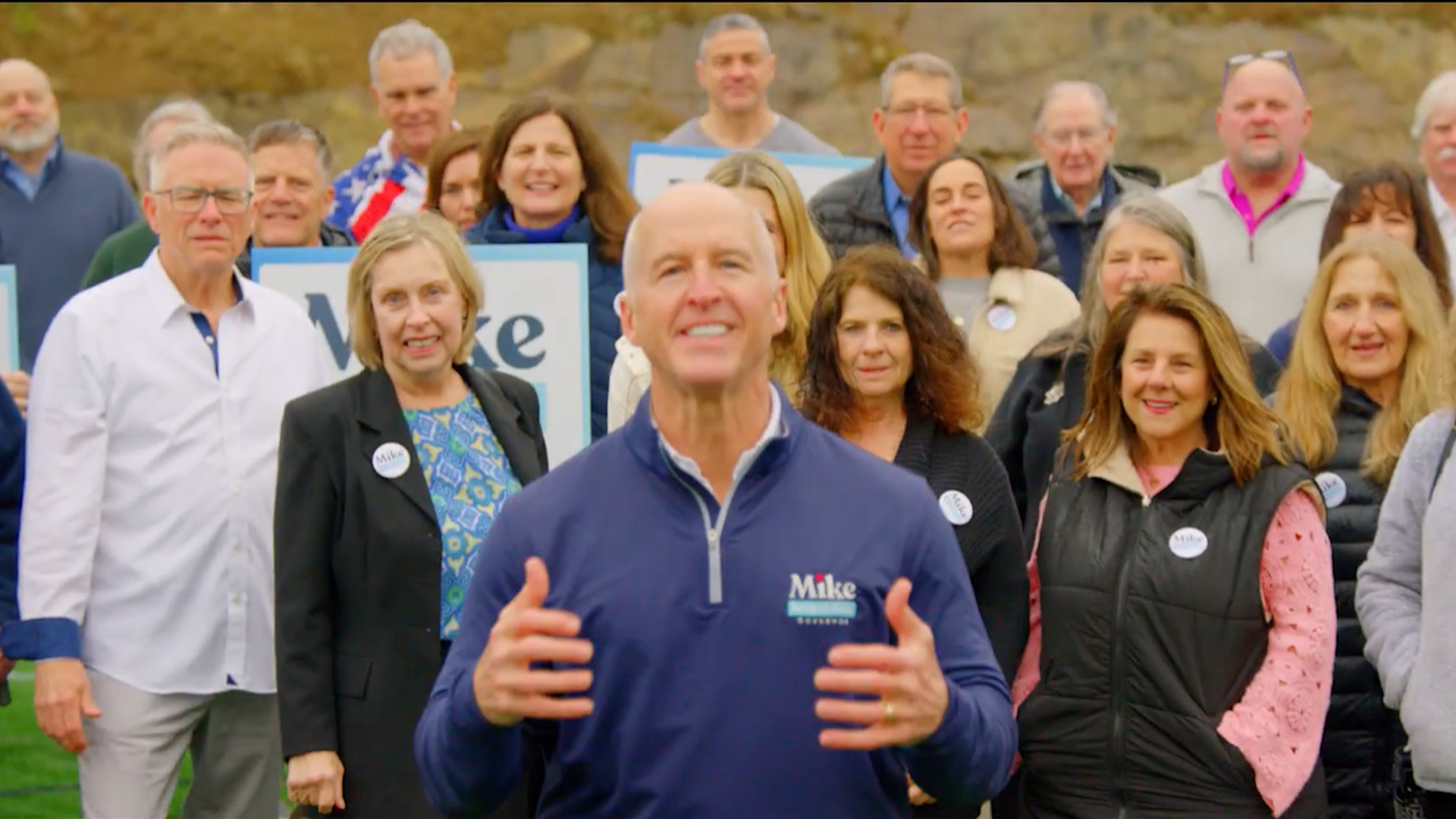 Large group of adults outdoors on a field, gathered around a smiling man in a blue pullover at the front who gestures toward the camera; campaign signs and Mike stickers visible behind them.