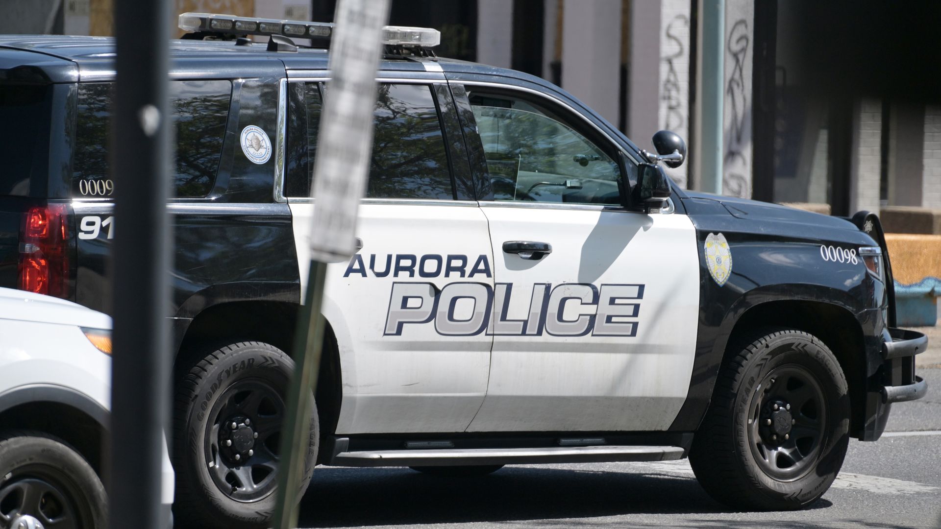 Side view of a dark blue and white Aurora Police SUV with a roof light bar, bold POLICE lettering, a badge sticker on the rear window, and an urban backdrop with graffiti.
