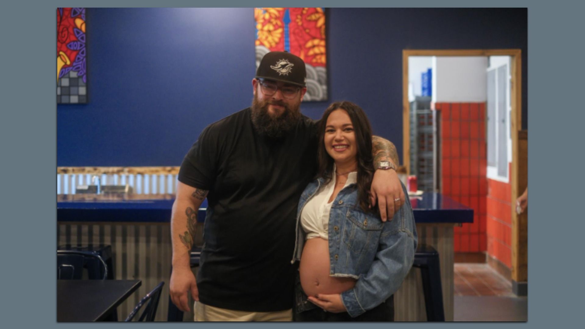 Smiling couple Jeff and Laura Bud  indoors with blue wall. Man wears black shirt and hat, woman shows pregnant belly, wearing denim jacket and white top. Red tiles visible through doorway.