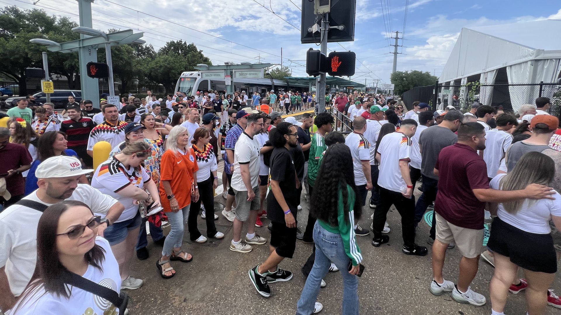 Crowd of people walking on a city street during an event; many wear white shirts and sports jerseys. Overhead tram lines, a pedestrian signal showing a red hand, and a tram in the background.