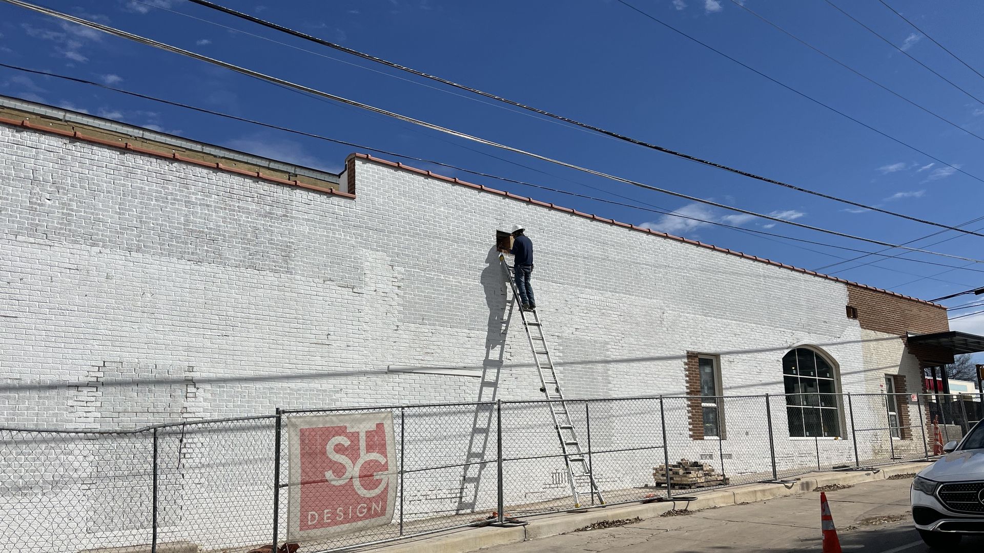 Person on ladder painting a large white brick wall under a clear blue sky, behind a chain-link fence with an "SG Design" sign, near a street with an orange traffic cone and a white car.
