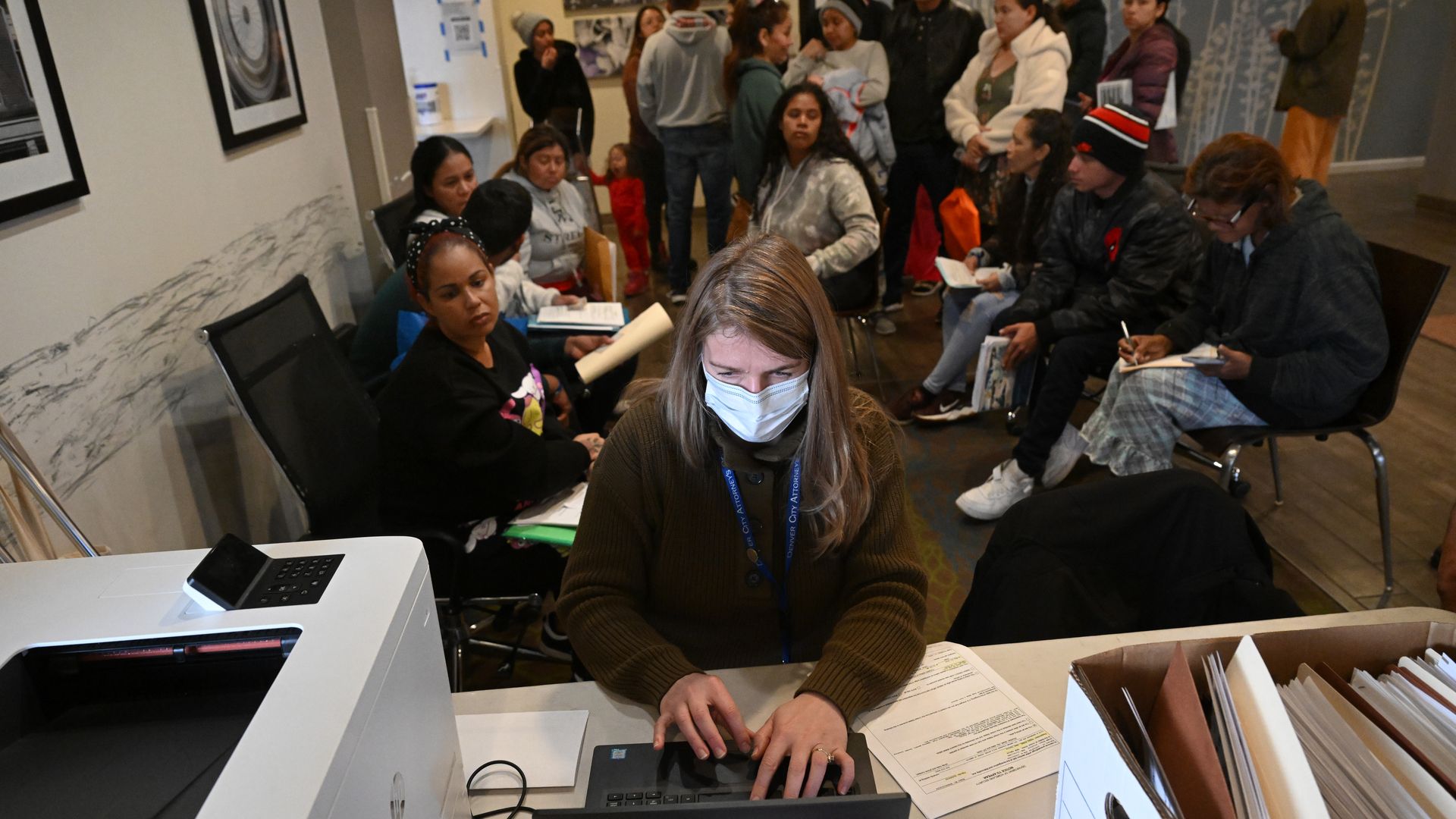 A woman sits at a computer with a dozen migrants standing and sitting behind her.