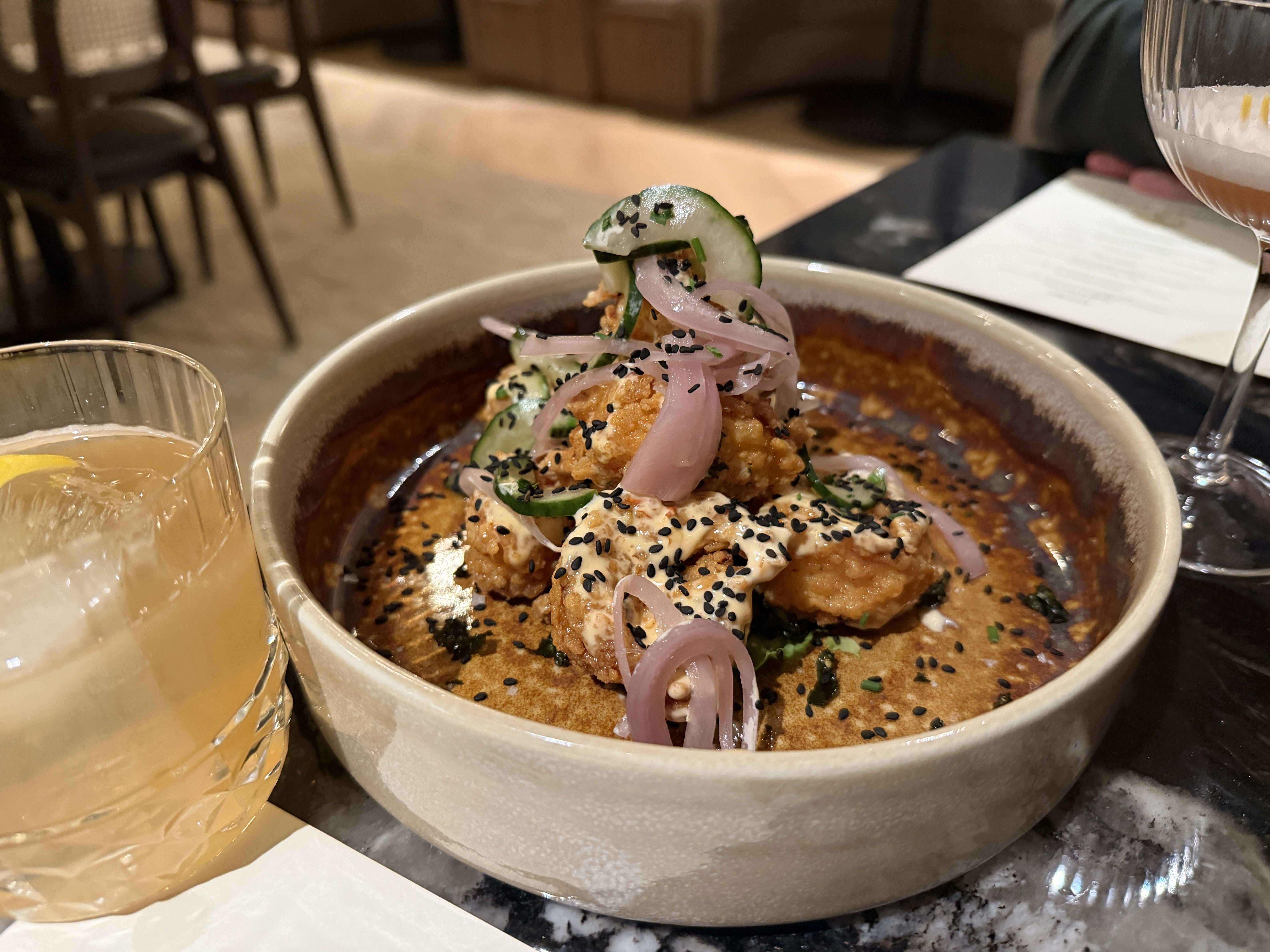 Brown ceramic bowl with fried cauliflower topped with pickled red onions, cucumber slices, black sesame seeds, and a creamy sauce, next to a glass of iced lemon drink on a restaurant table.