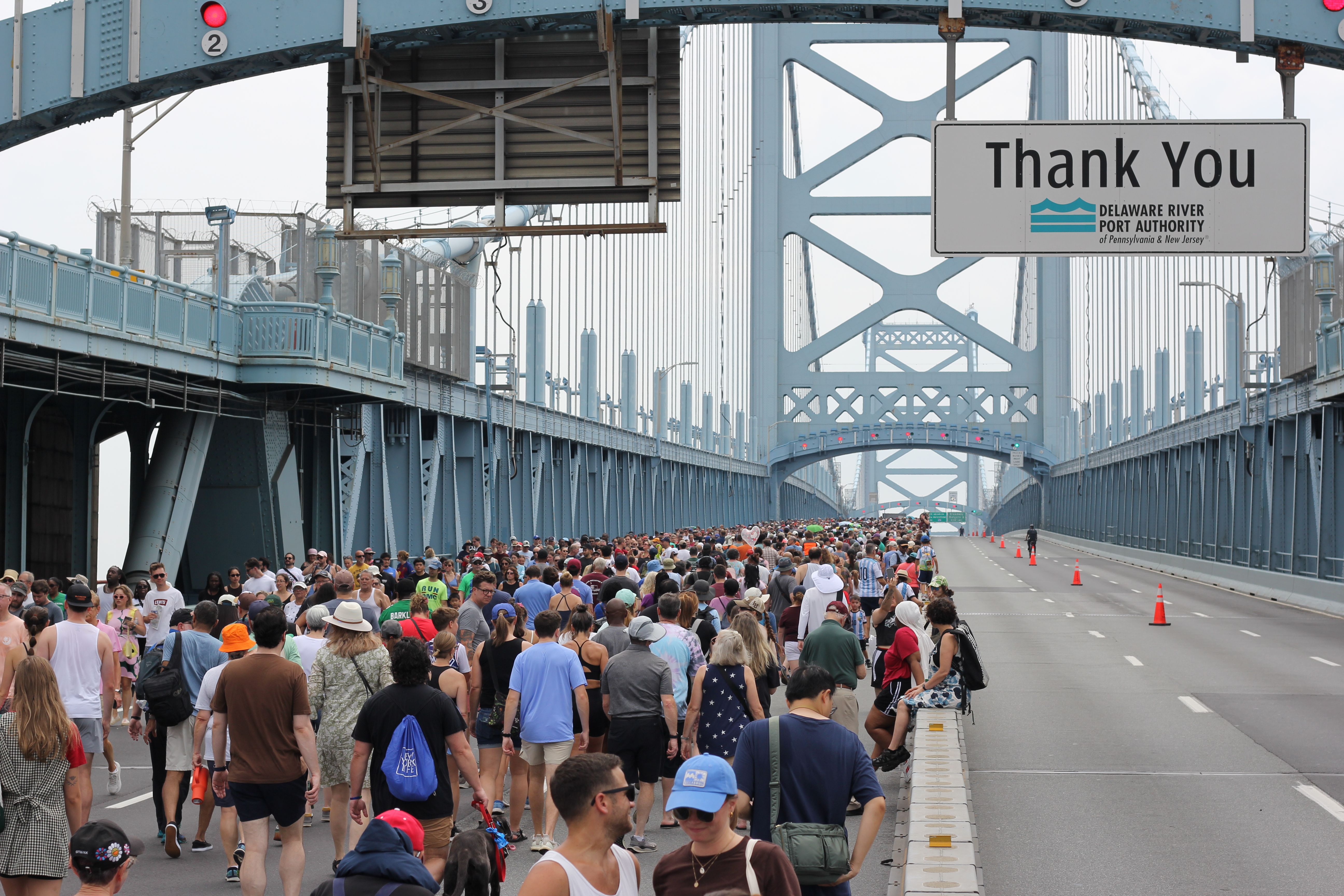 A dense crowd of people walks on a blue steel suspension bridge, with a large Thank You sign from the Delaware River Port Authority hanging over the roadway; orange cones line the right lane.