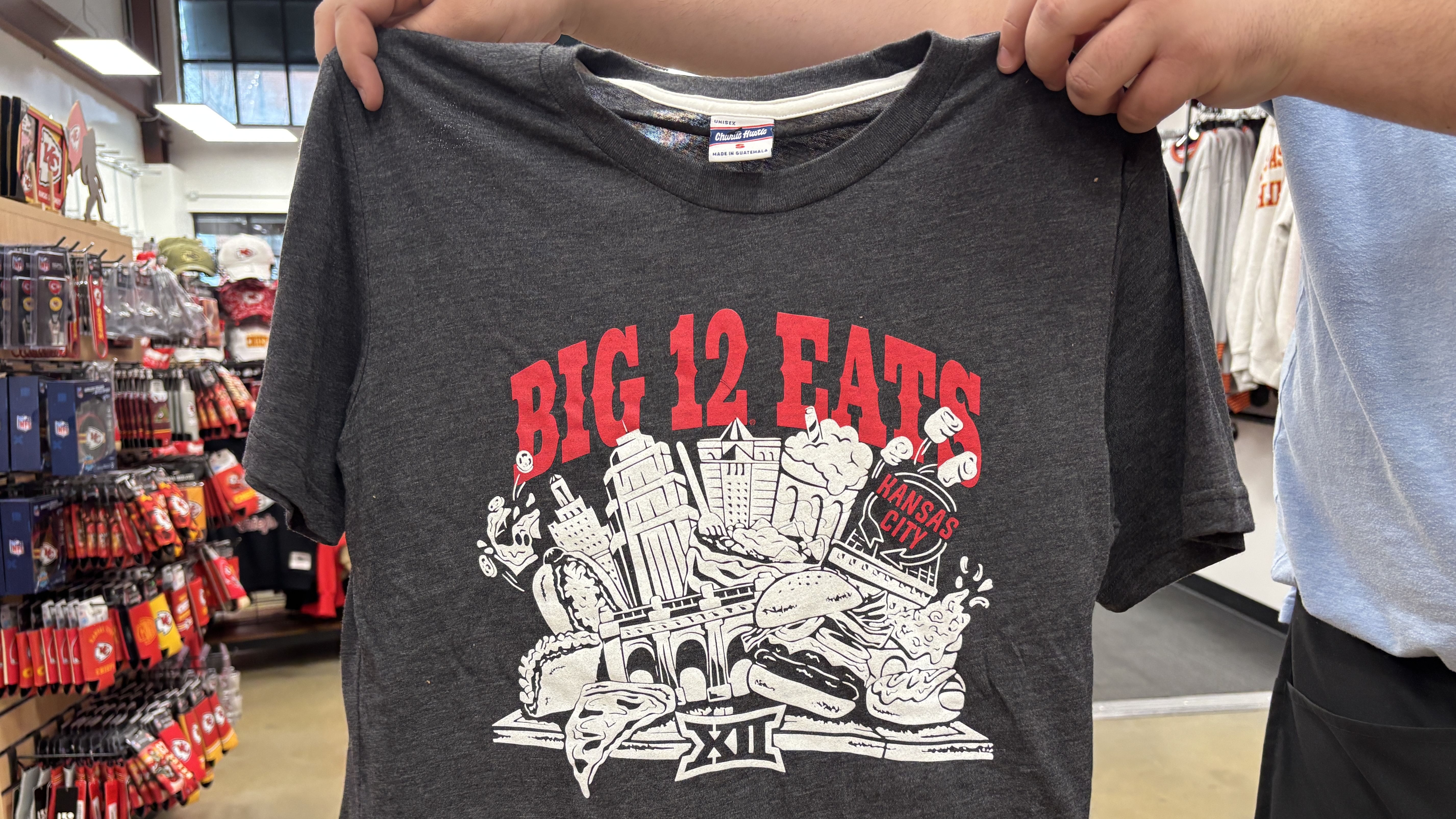 Gray T-shirt held up in a store, featuring red text "BIG 12 EATS" and white graphics of Kansas City landmarks and various foods, including pizza, burgers, and drinks.