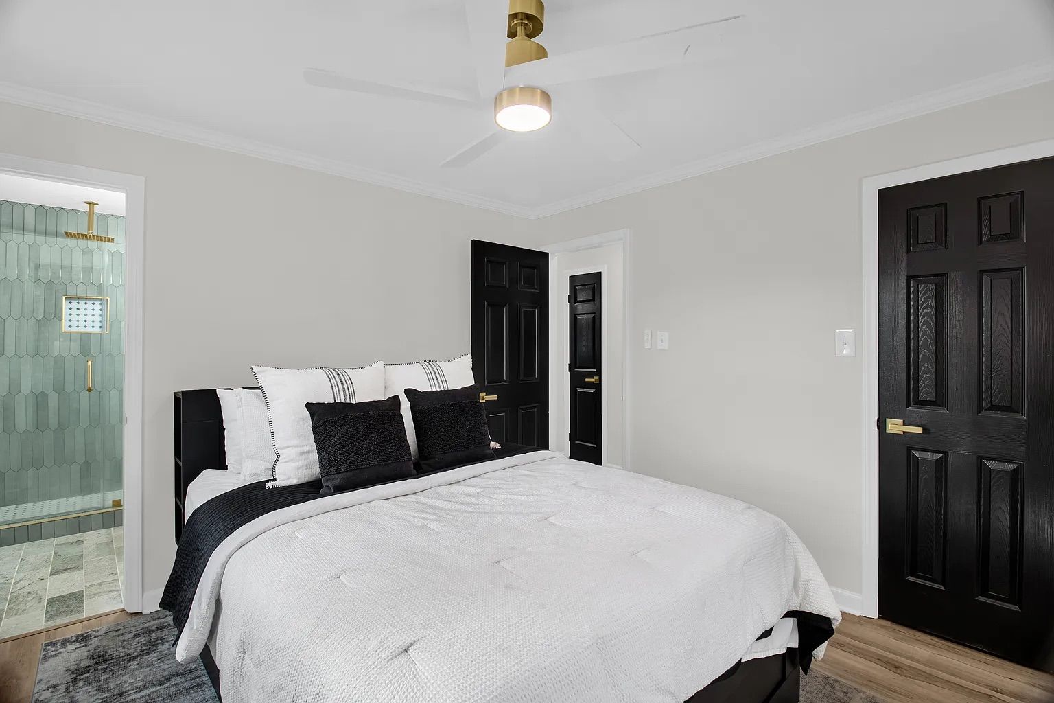 Modern bedroom with white walls, black doors, a bed with white and black pillows and bedding, a ceiling fan with a gold base, and a glimpse of a bathroom with green tiles and gold fixtures.