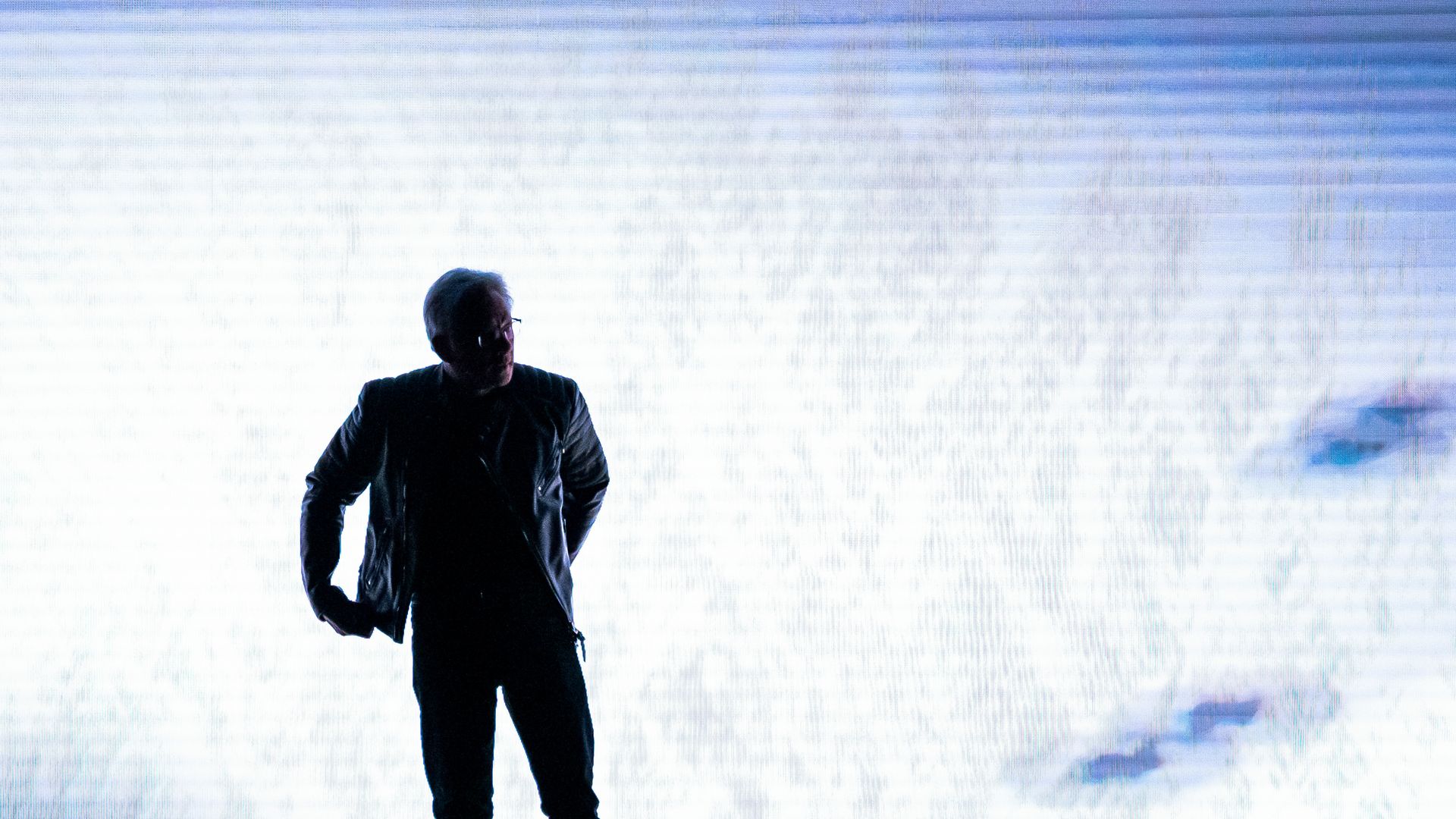 A person in a leather jacket stands in front of a screen with clouds on it