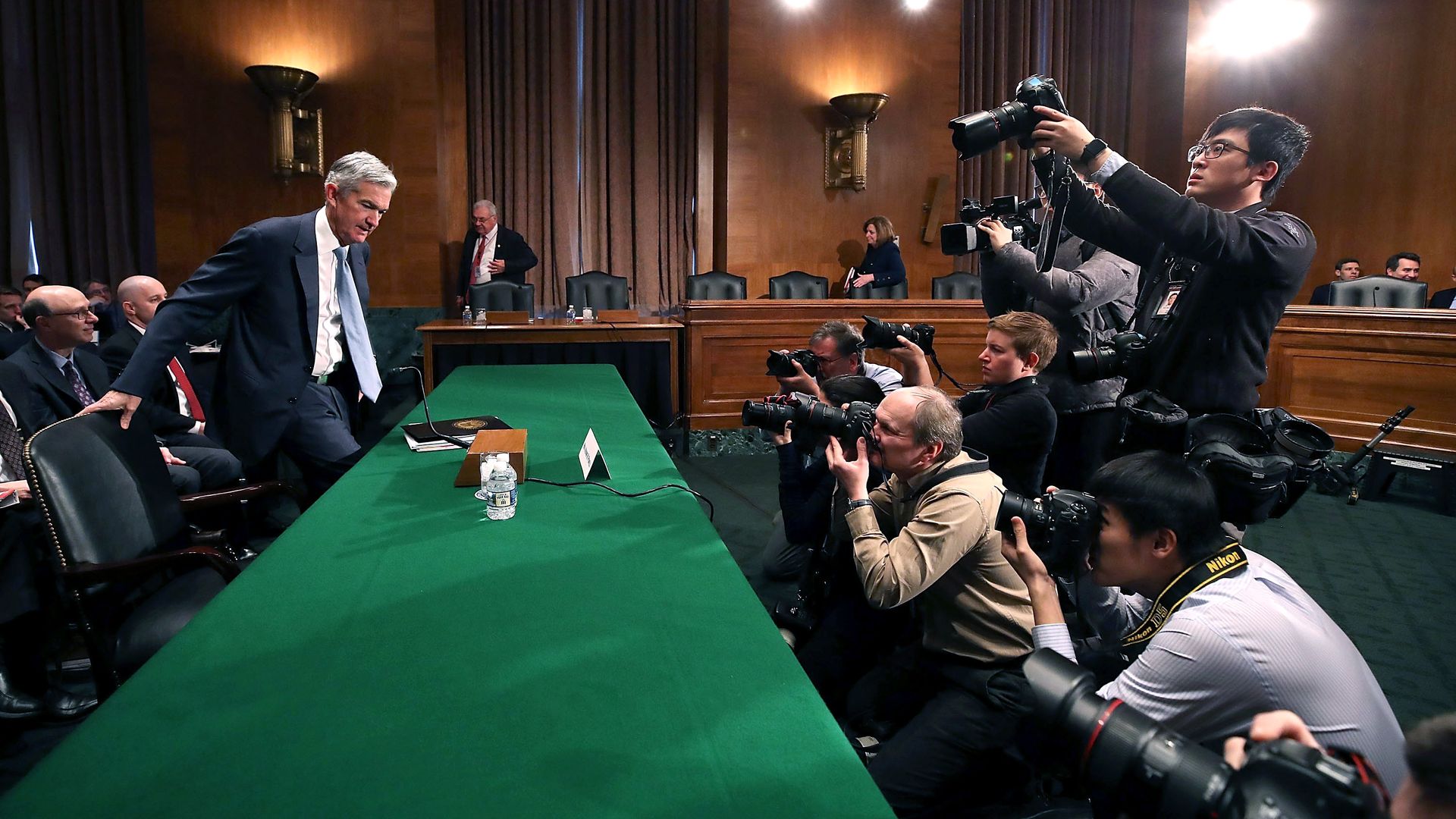 Fed Chair Jerome Powell sits down at a large green table as photographers snap pictures.