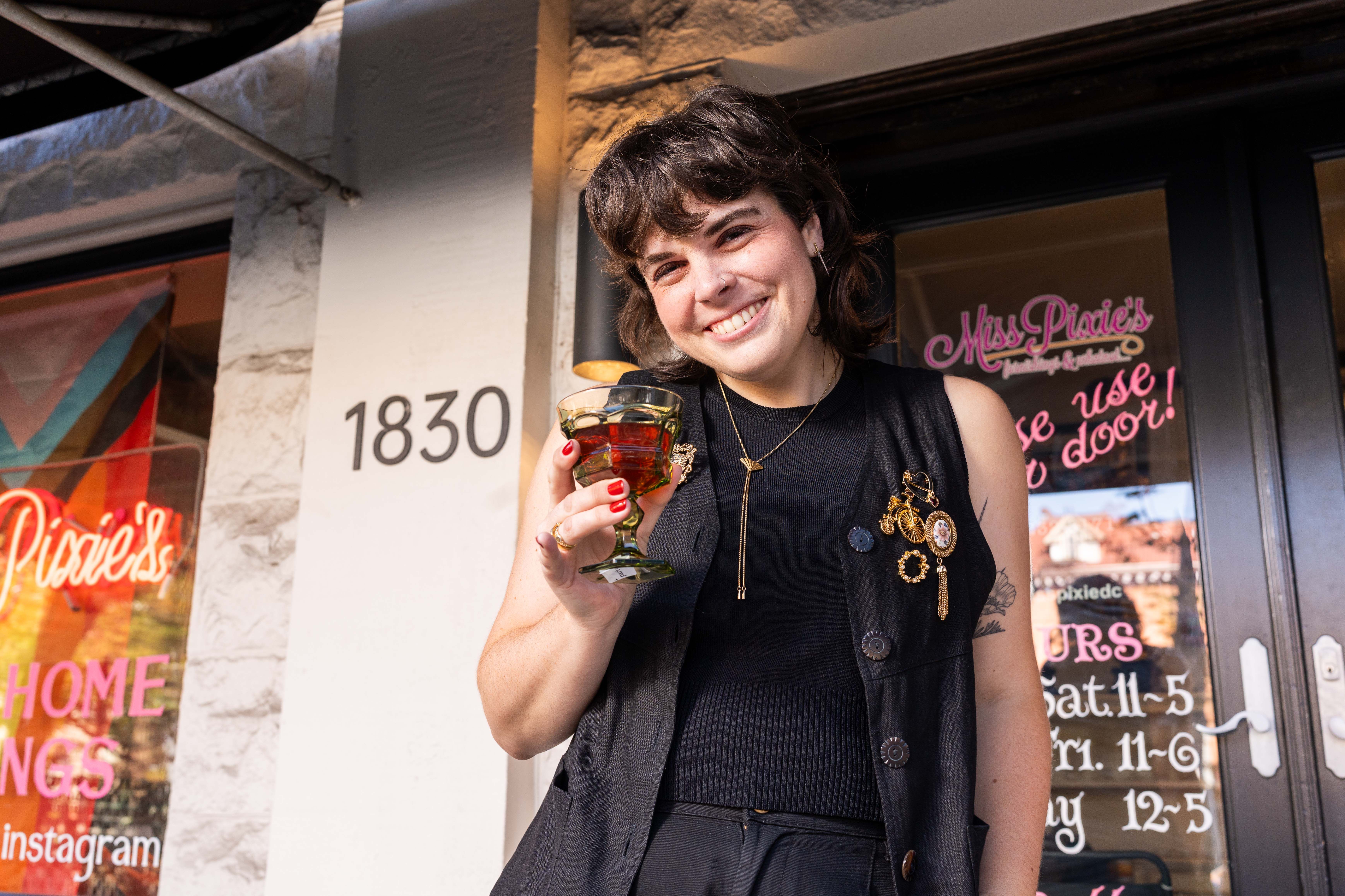 Smiling person with short dark hair holding a drink in a green glass, wearing a black sleeveless outfit with decorative pins, standing outside a storefront.
