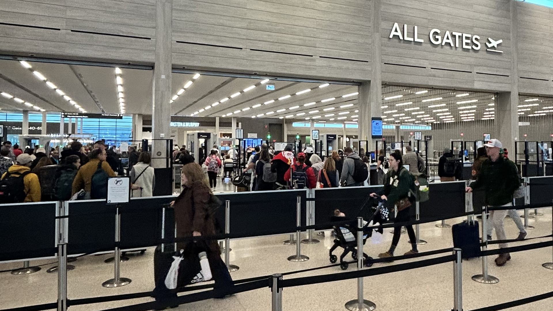 Crowded airport security checkpoint with travelers lined up, some pushing strollers and rolling suitcases. Large sign reading "ALL GATES" with airplane icon overhead.