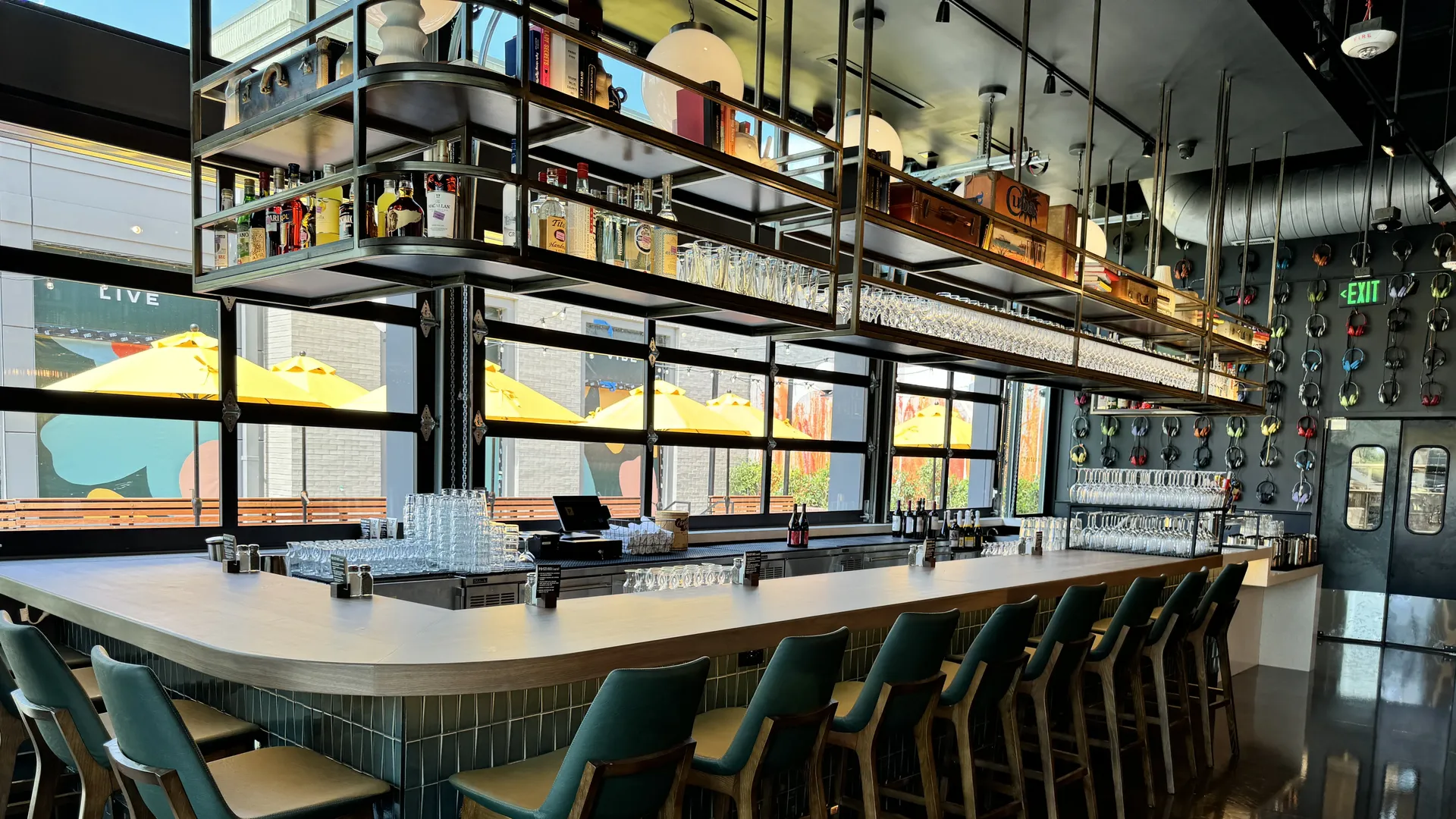 Modern bar interior with overhead metal shelves stocked with liquor bottles and glassware. A long tiled counter with teal stools, large windows, and yellow outdoor umbrellas visible outside; green exit sign nearby.