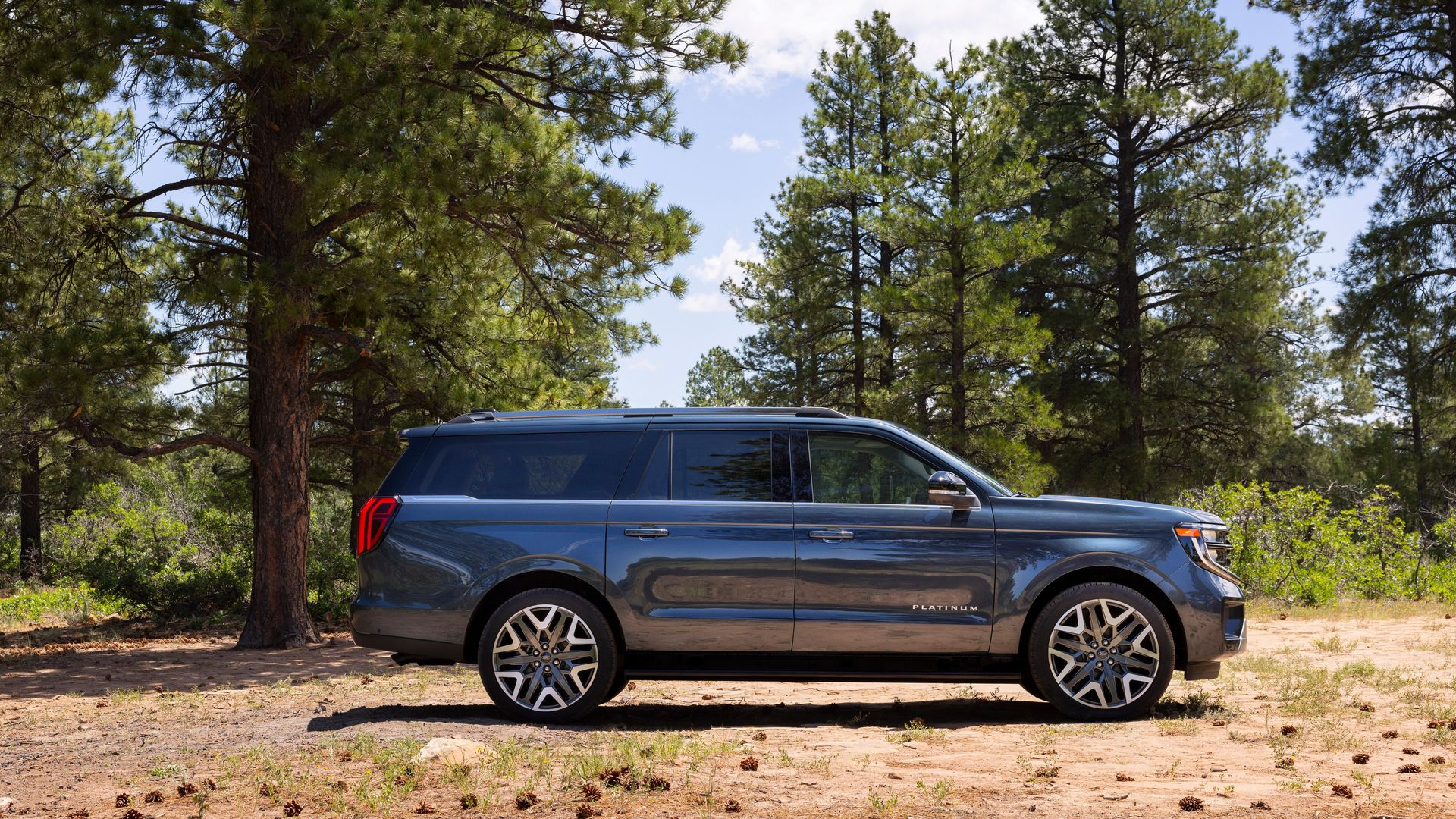 Side view of a dark gray 2025 Ford Expedition Platinum MAX SUV parked on a dirt patch surrounded by tall green pine trees under a partly cloudy blue sky.