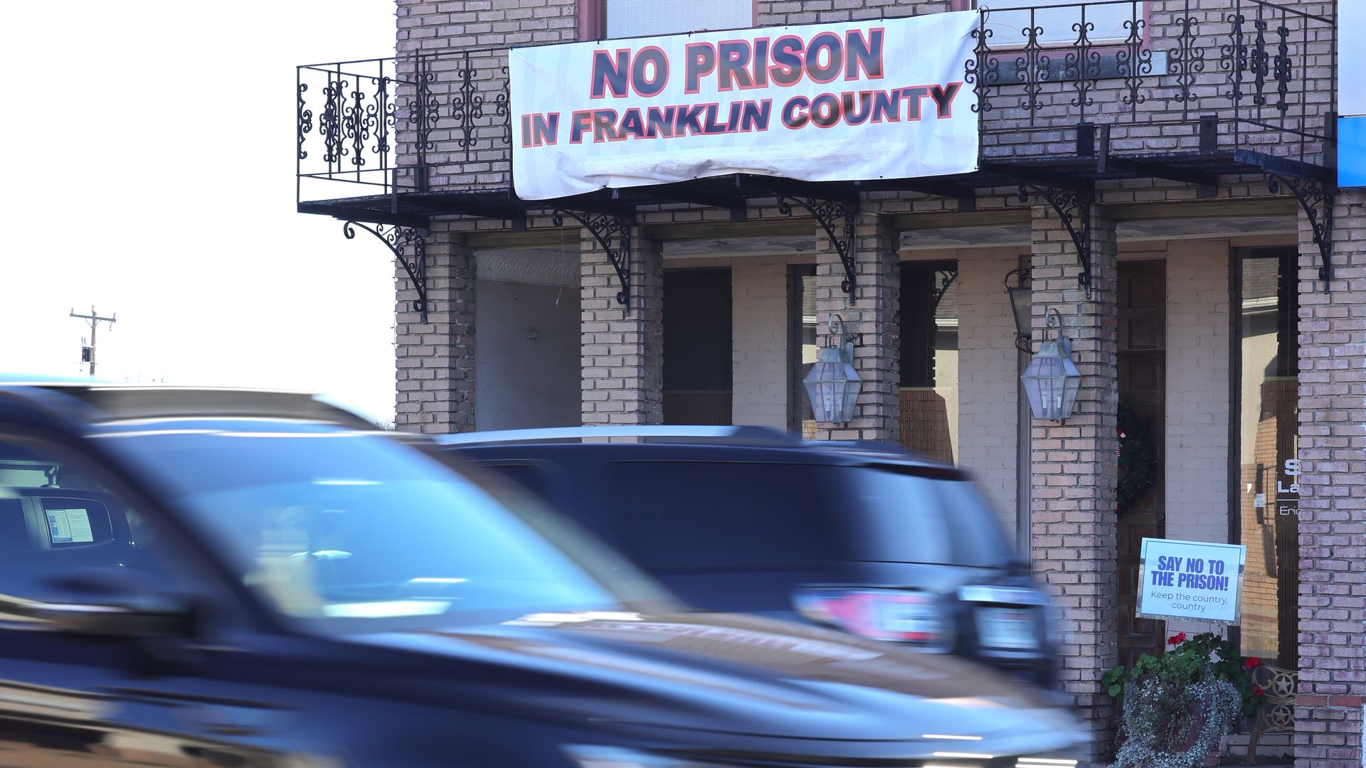 Building with a white banner stating "NO PRISON IN FRANKLIN COUNTY" in red text, and a small sign saying "SAY NO TO THE PRISON!" near a flower pot; blurred black car passing by on street.