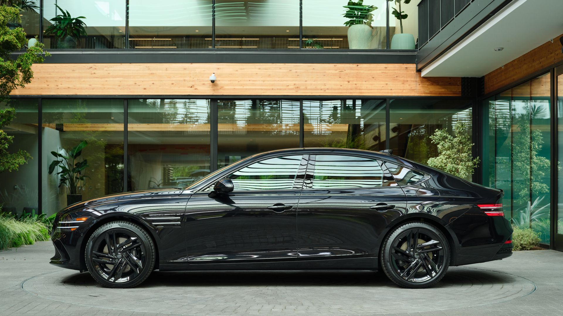 Shiny black luxury sedan parked before a modern glass-and-wood building with large windows, greenery, and a second-floor glass railing.