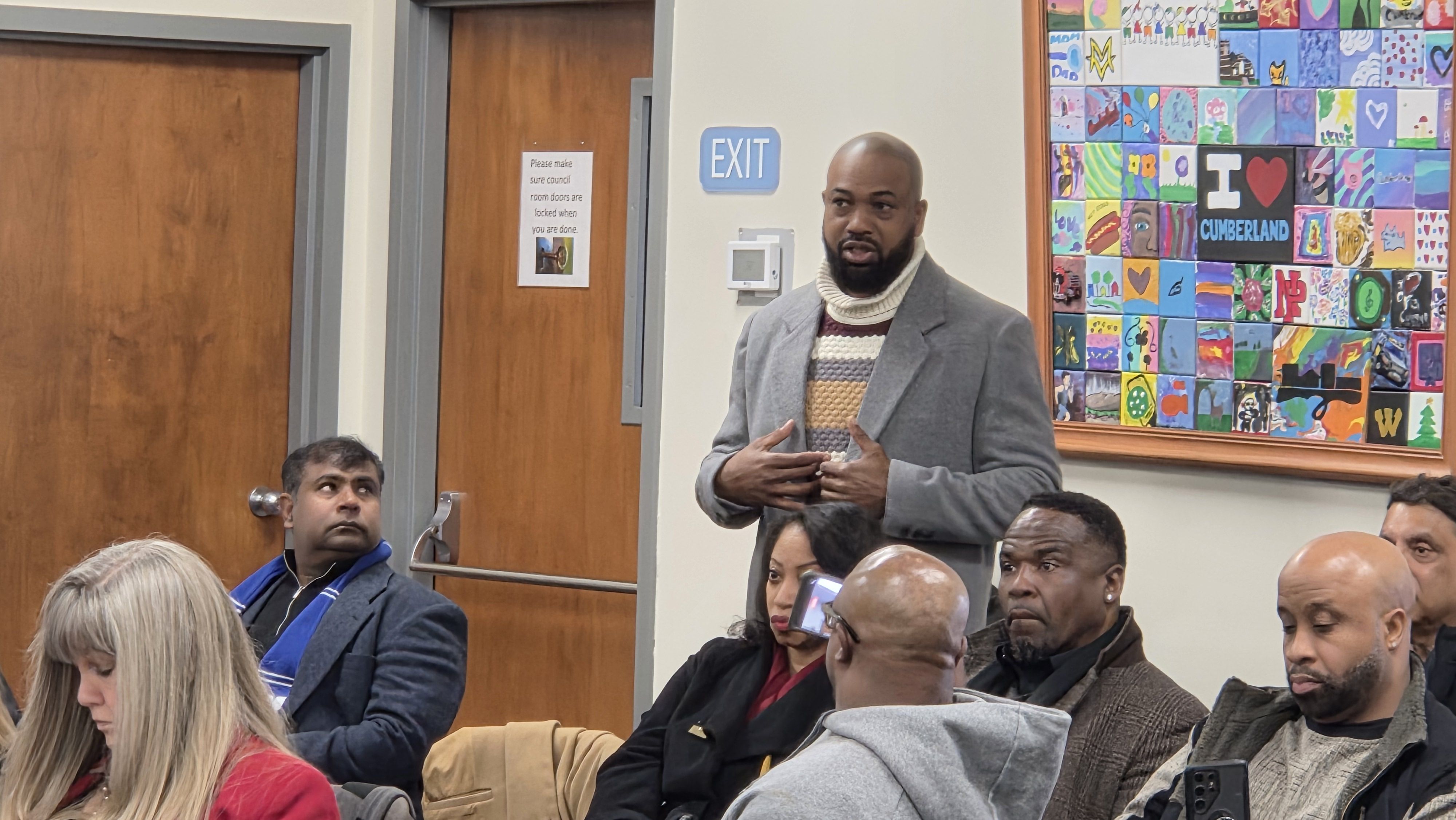 A man in a gray coat and striped turtleneck sweater speaks to a seated group in a room with wooden doors and a colorful "I love Cumberland" tile mural on the wall.