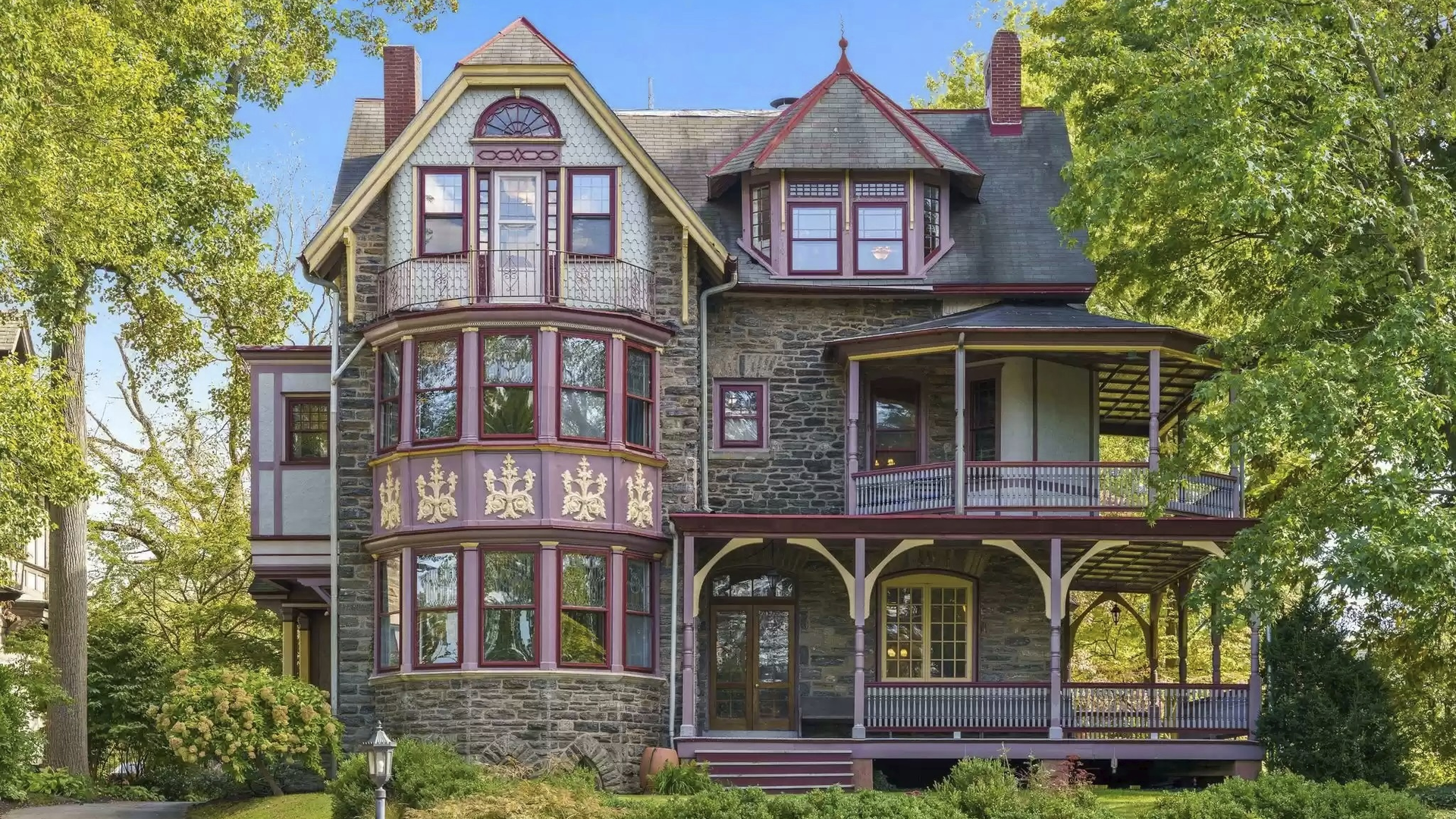 Victorian stone house with purple trim, ornate bay windows, and a wraparound porch, set among lush green trees under a bright blue sky.