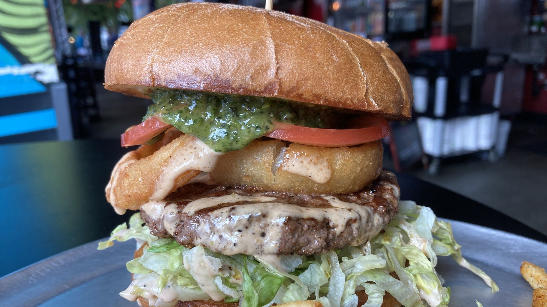 A hamburger stacked with shredded lettuce on the bottom, then a brown patty, a tan fried onion ring, a red tomato and a thick drip of green sauce falling out from under the top bun.