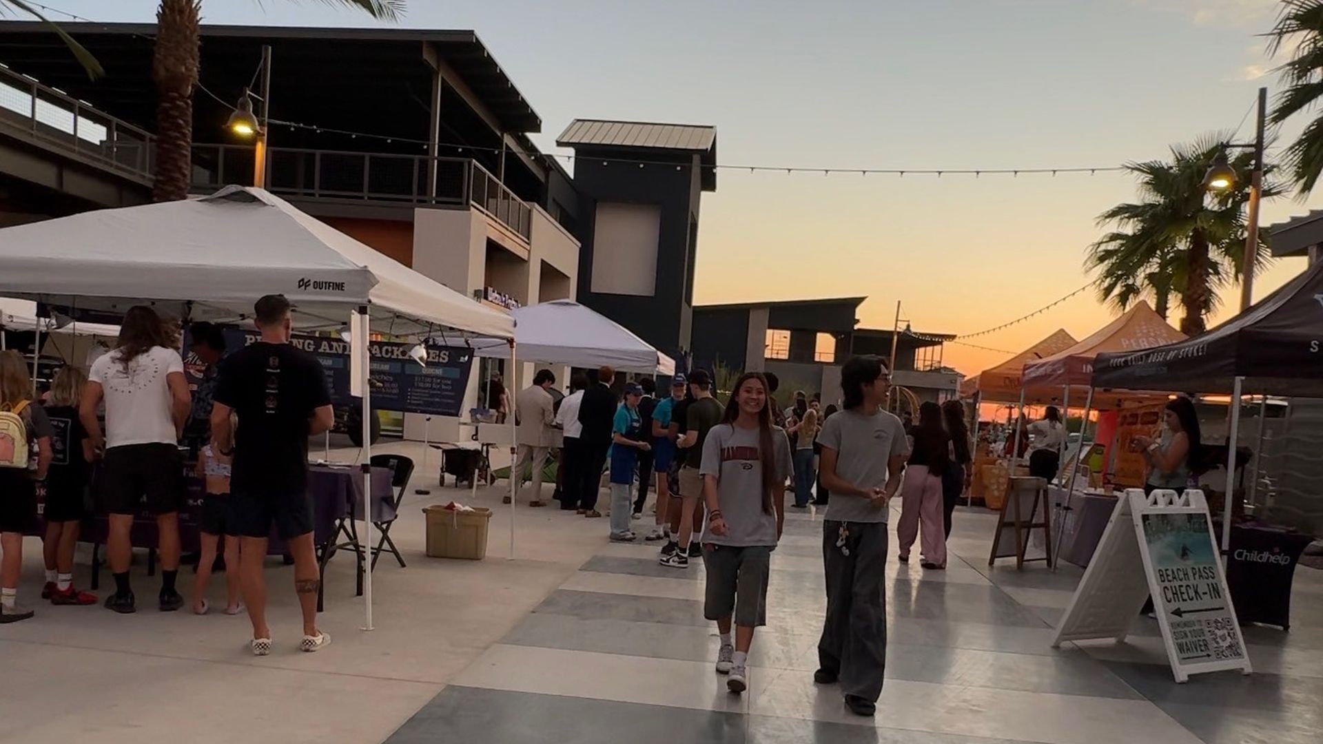 Sunset outdoor market with white tents and people walking along a tiled plaza; palm trees, string lights, and modern buildings in the background under a warm orange sky.
