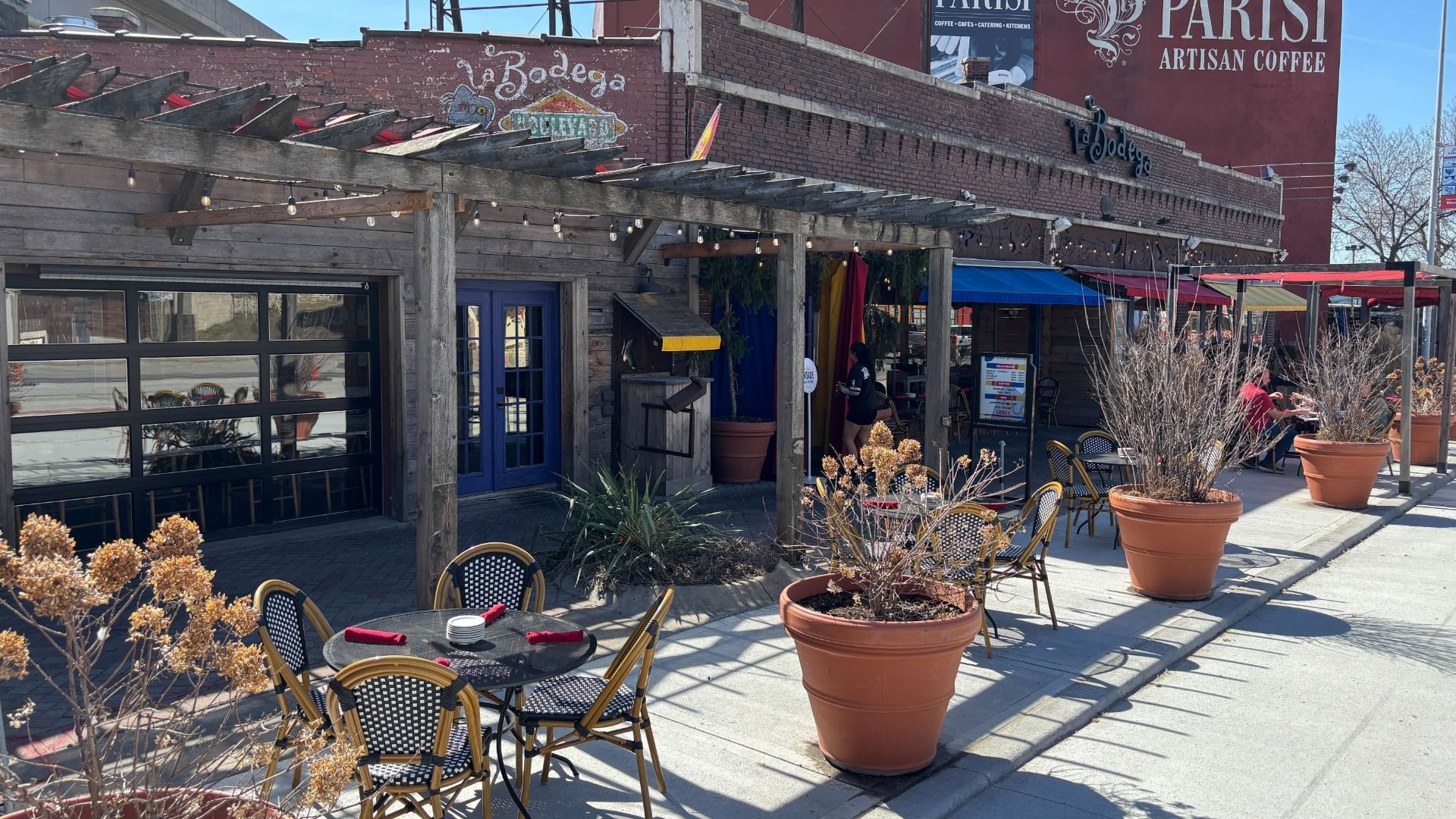 Outdoor street café under a wooden pergola with string lights. Round tables with black-and-white chairs, red napkins, and large potted plants. Blue door and brick façade with a PARISI sign.