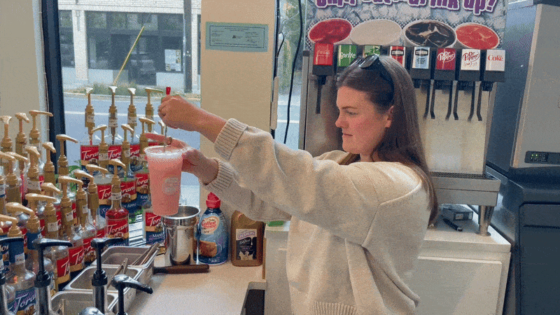 A woman in a cream sweater uses a red pump to add syrup to a pink soda in a convenience store. She stands in front of syrup bottles and a soda fountain, concentrating on the drink preparation.