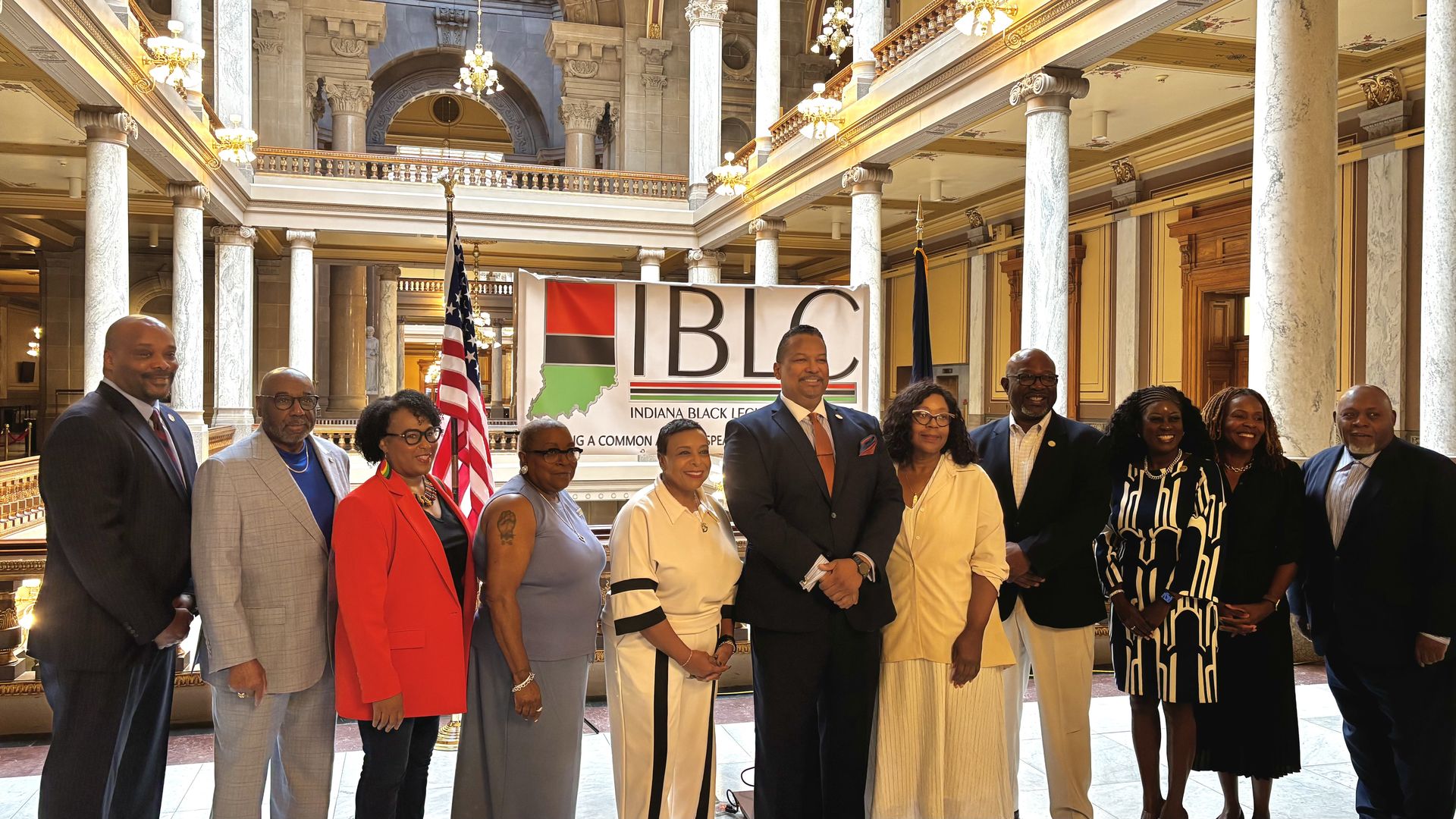 A diverse group of men and women stands in a grand marble atrium with tall columns and chandeliers, behind them a large banner displaying IBC and Indiana Black text, flanked by American flags.
