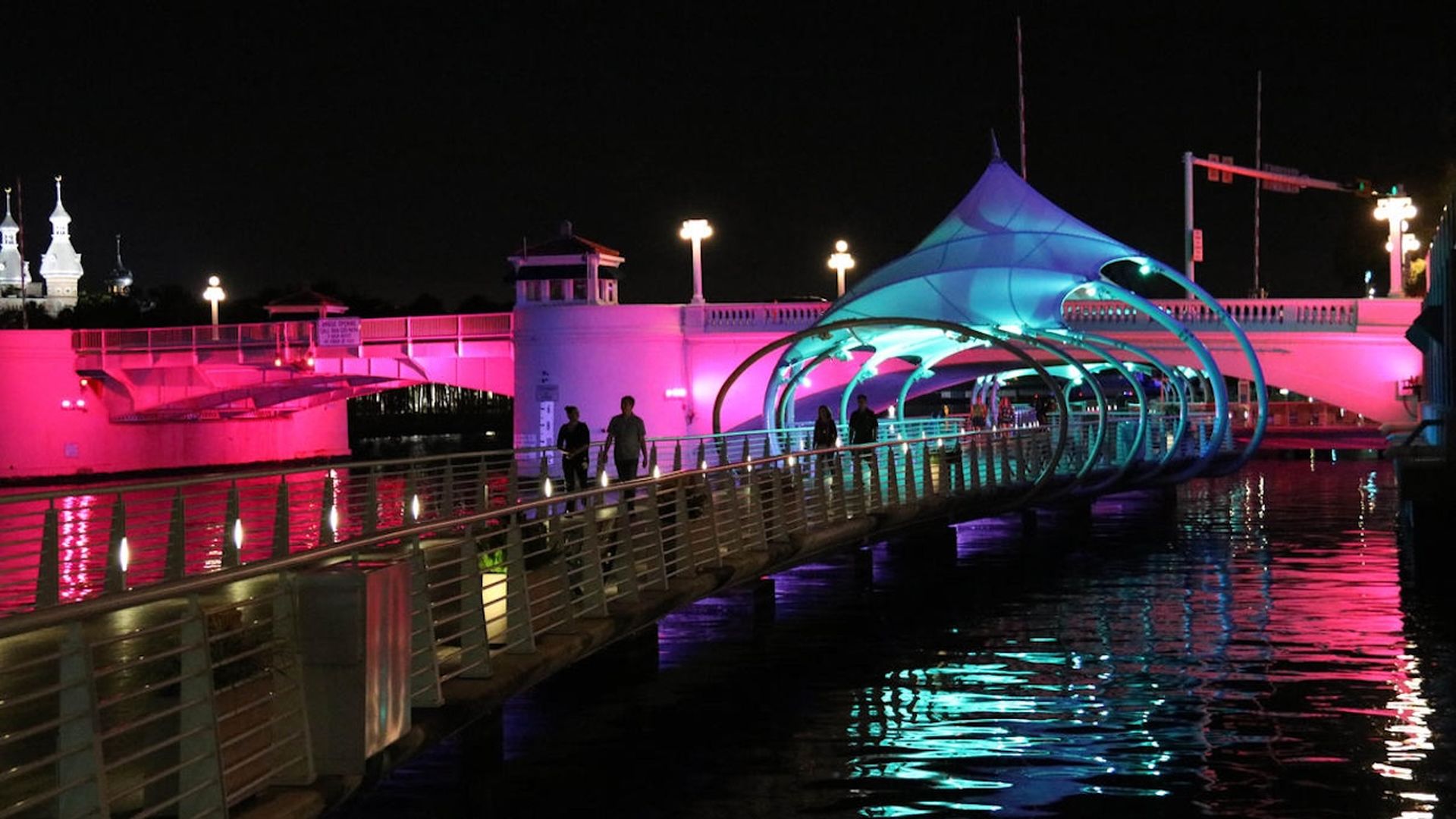 A footbridge with blue and purple lights arches over water at night, with a larger bridge lit in pink behind it. Several people walk along the footbridge.