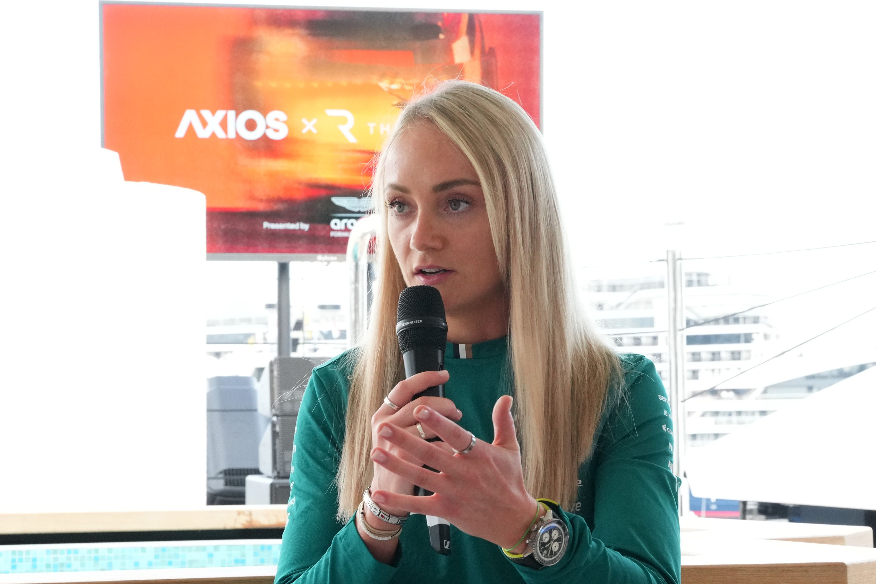 Aston Martin Aramco F1 Team head of F1 Academy and driver ambassador Jessica Hawkins in a green shirt speaks into a handheld black microphone, with an orange banner in the background reading "Axios x The Race Media."