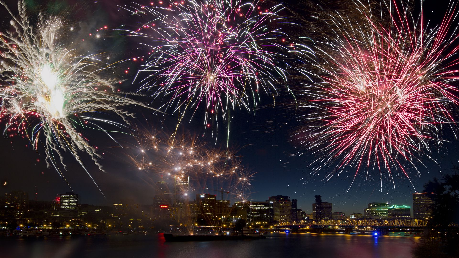 A vibrant display of fireworks in the night sky over a city skyline. The fireworks burst into a variety of colors, including red, pink, white, and green, illuminating the city below. Buildings along the waterfront are lit up, and their lights are reflected in the calm water of the river.