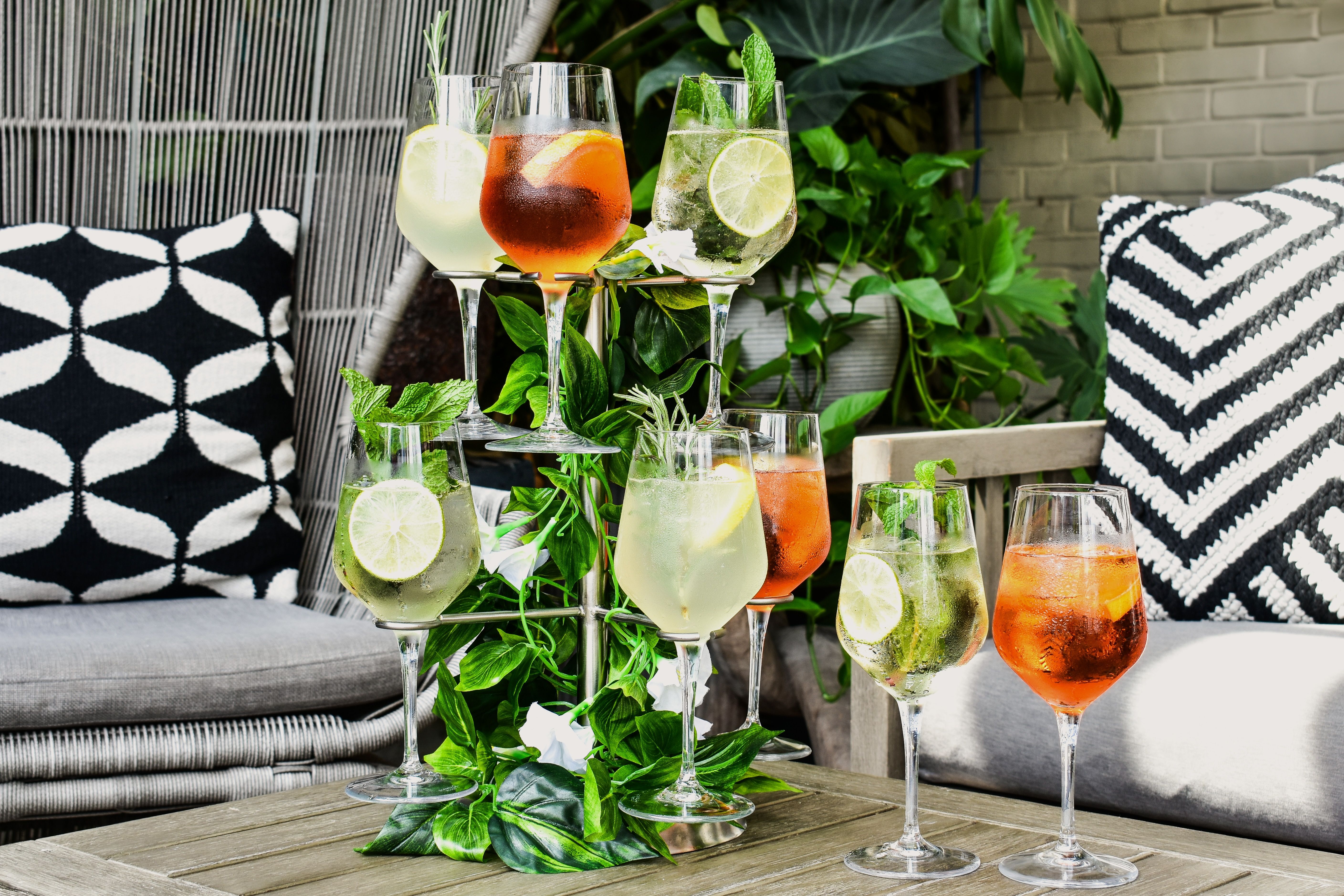 Three bright cocktails on a tiered glass stand, garnished with lime wedges and mint, set in an outdoor lounge with green plants and black and white geometric cushions.