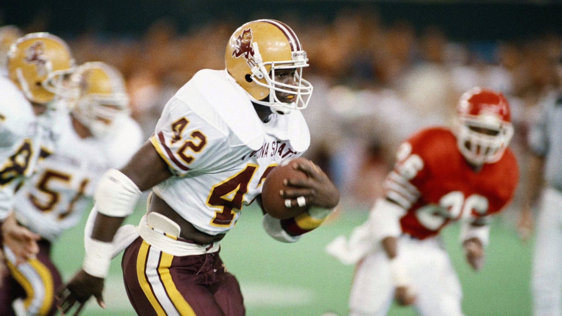 An Arizona State football player wearing No. 42, in a white jersey with maroon pants and a yellow helmet, carries the ball as a player in a red jersey gives chase in the background.