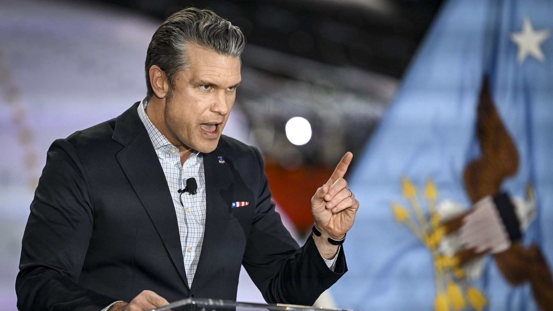 Man in dark blazer and checked shirt speaking passionately with raised finger, standing in front of a blue flag featuring an eagle and a white star.