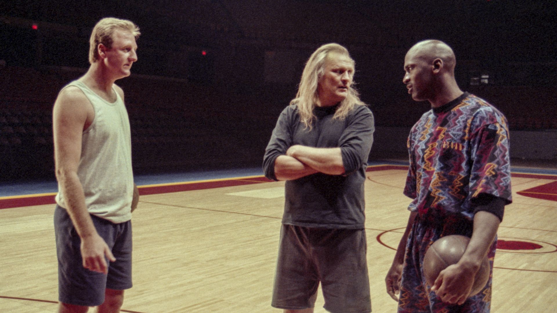 A person in a white tank top talks to a person in a colorful short-sleeved shirt holding a basketball on a basketball court. between them stands someone with long hair with his arms crossed