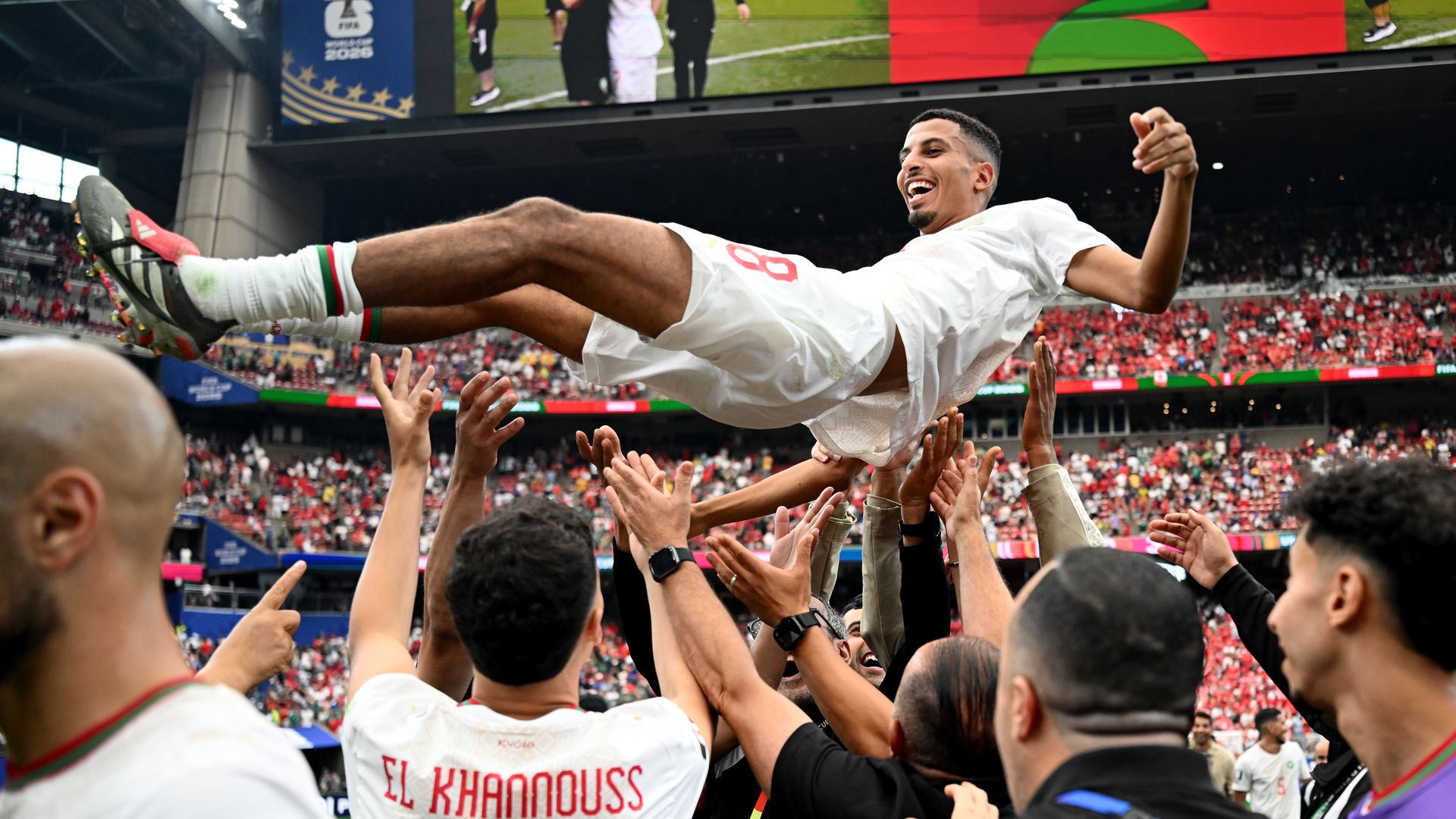 Soccer player in a white kit is hoisted by cheering fans in a packed stadium. His jersey reads El Khannouss as he smiles, with a large screen showing Morocco in the background.