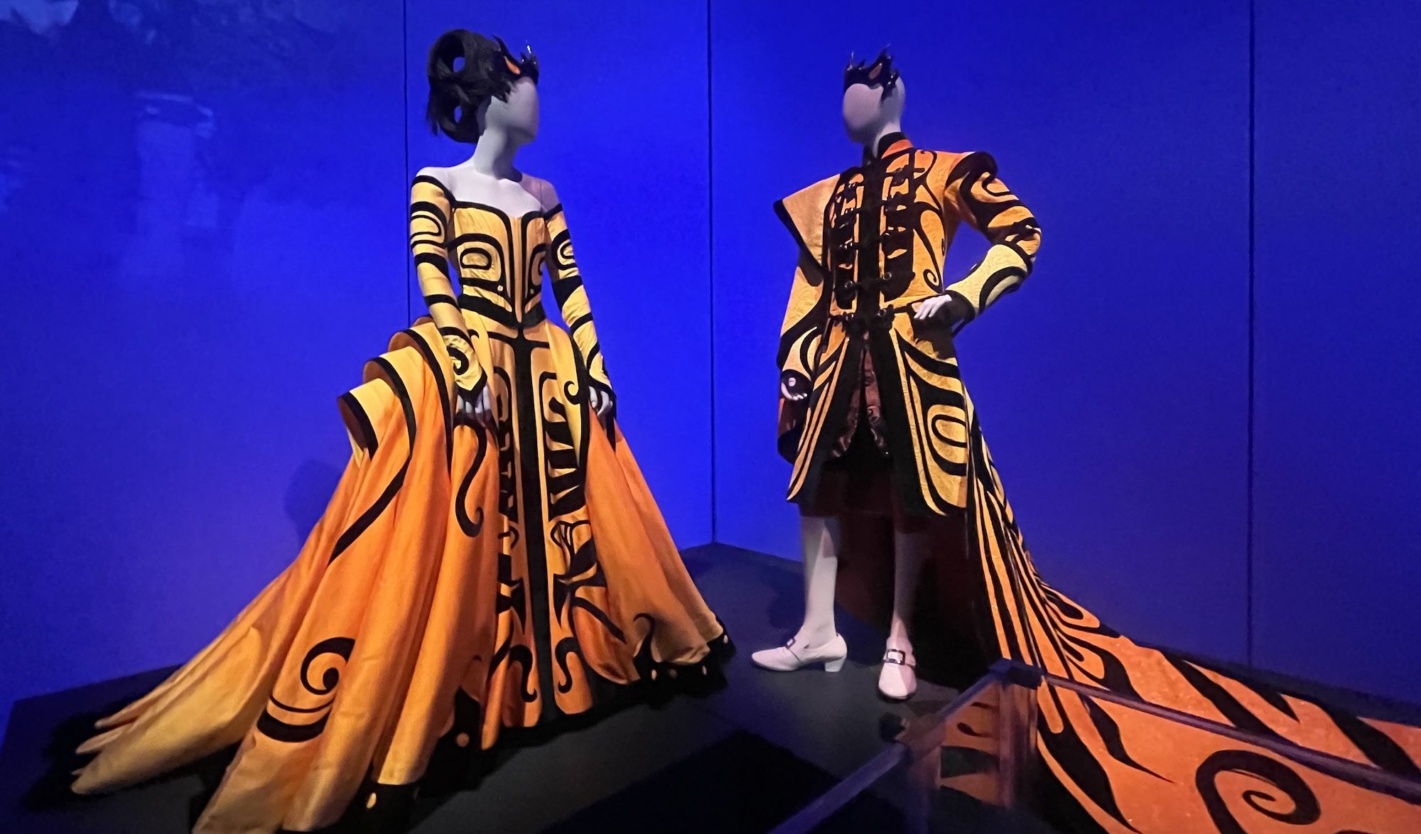 Two mannequins dressed in bright orange gowns with black tribal patterns, set against a deep blue background, evoking a dramatic and artistic fashion display.
