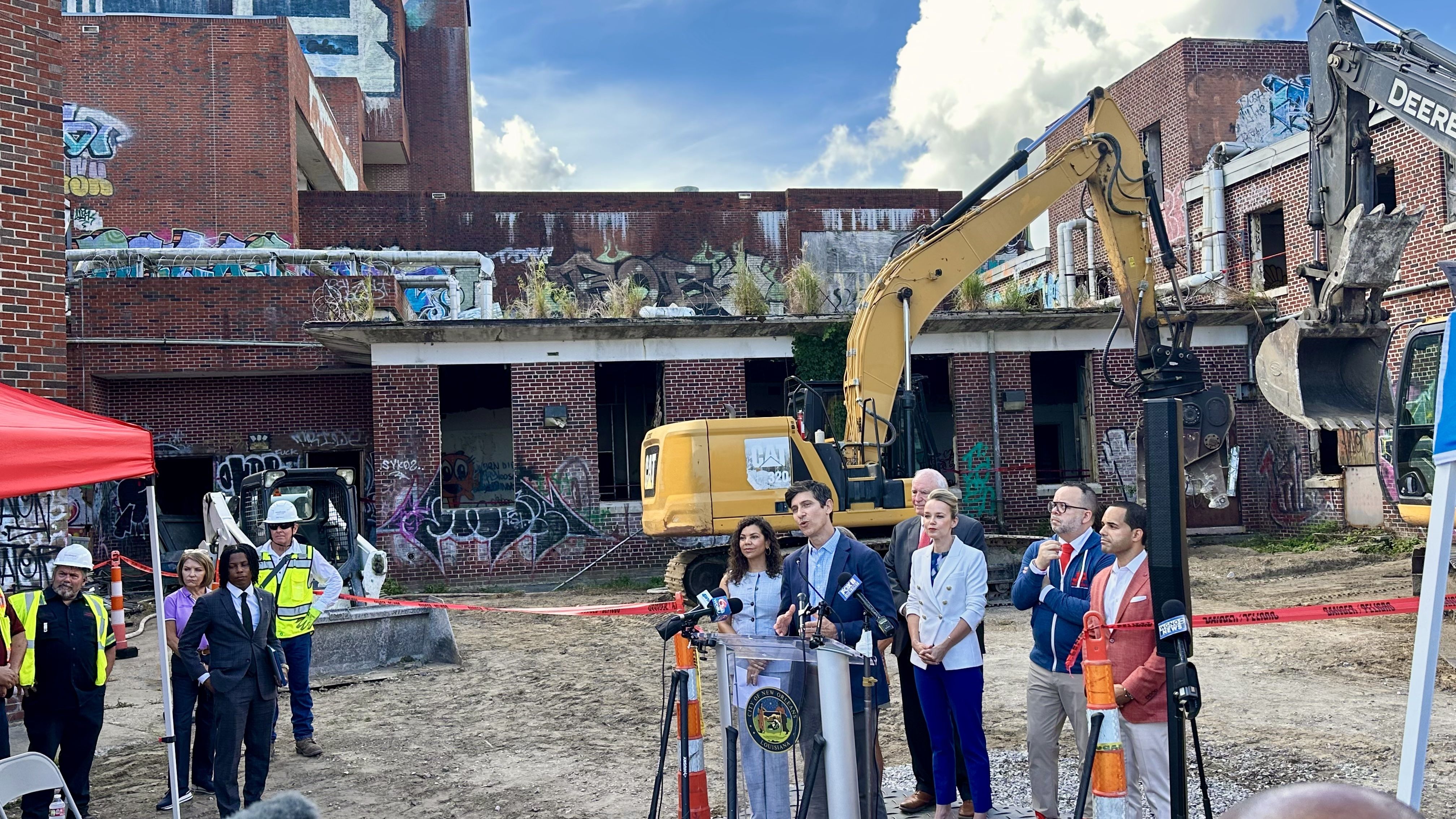 Construction site with a yellow excavator in front of graffiti-covered brick buildings. A group of officials and reporters stand behind red caution tape, microphones at a podium.