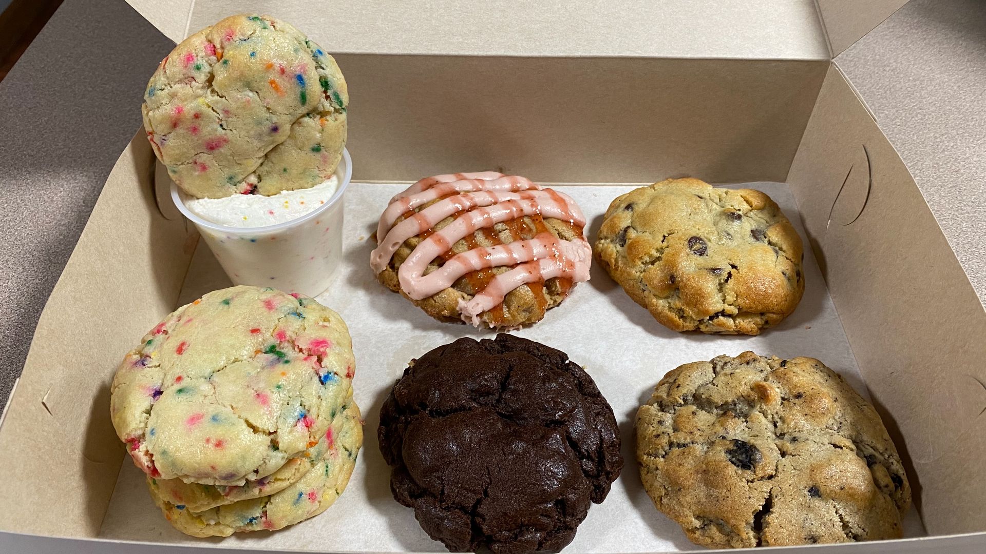 Four cookies in a box, plus a stack of sugar cookies with rainbow sprinkles and one inside a cup of frosting