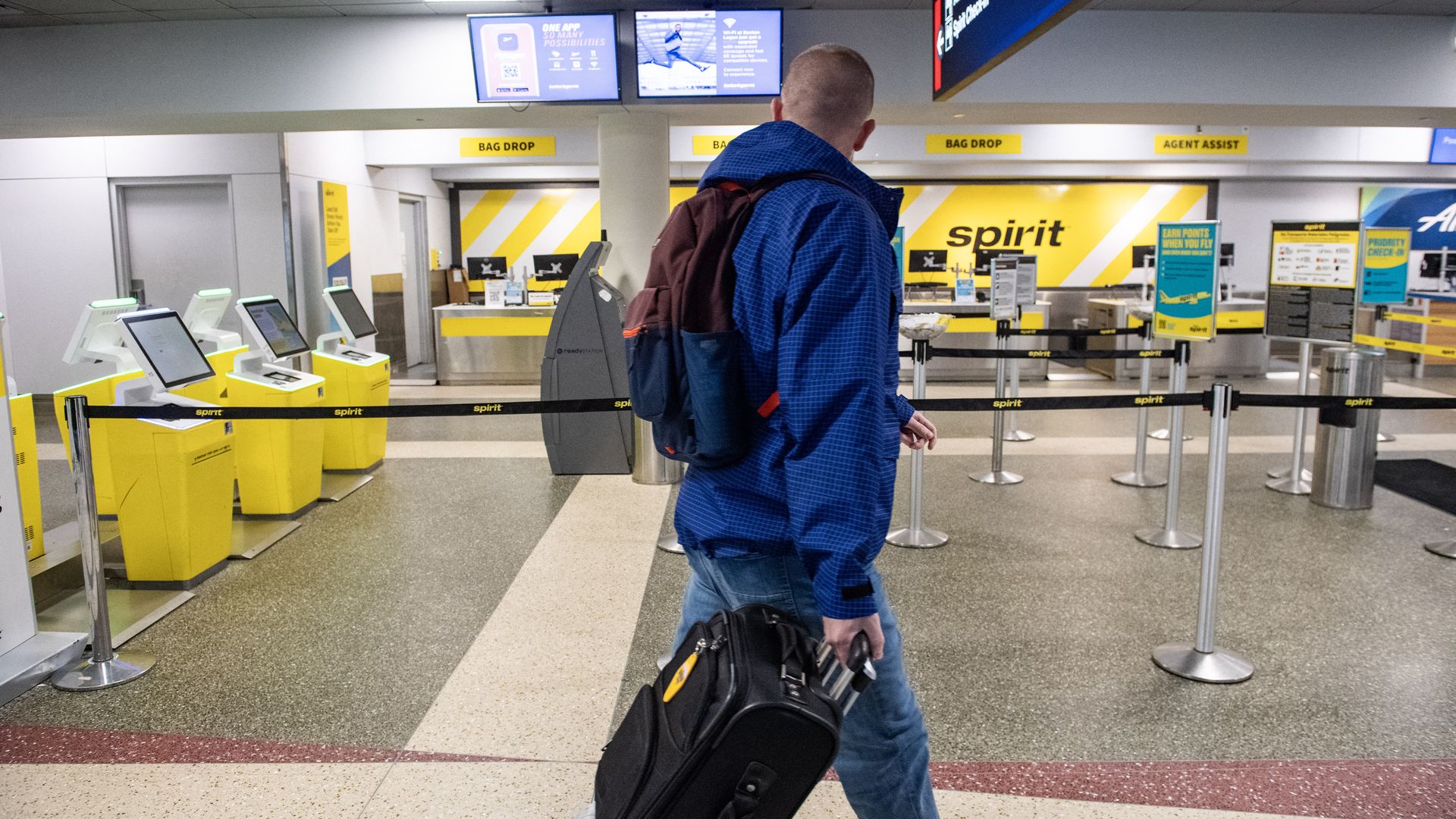 Back view of a man in a blue jacket and backpack pulling a black rolling suitcase through a Spirit Airlines check-in area with yellow counters, bag-drop signs, and kiosks.