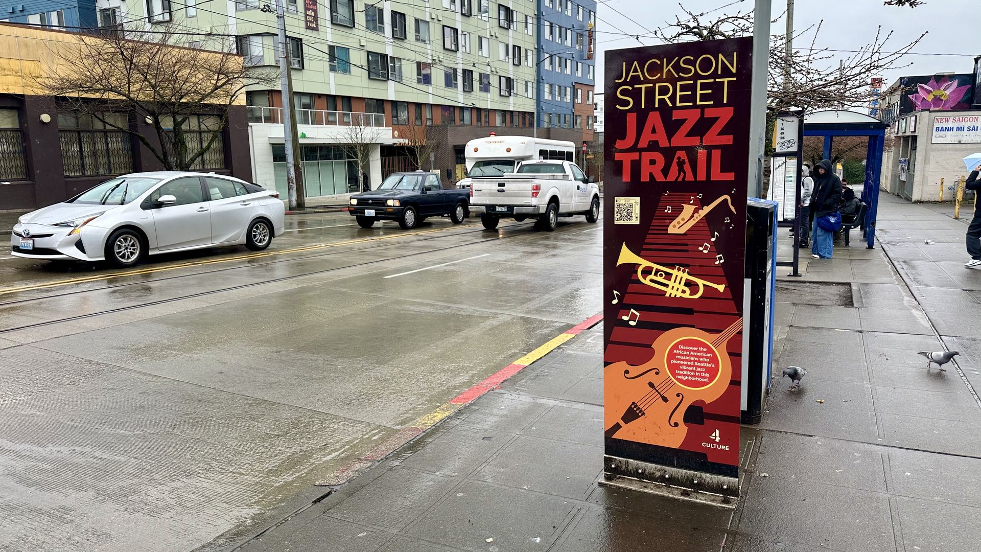 A pillar on a sidewalk that reads "Jackson Street Jazz Trail" near a bus stop and a street with cars driving on it.
