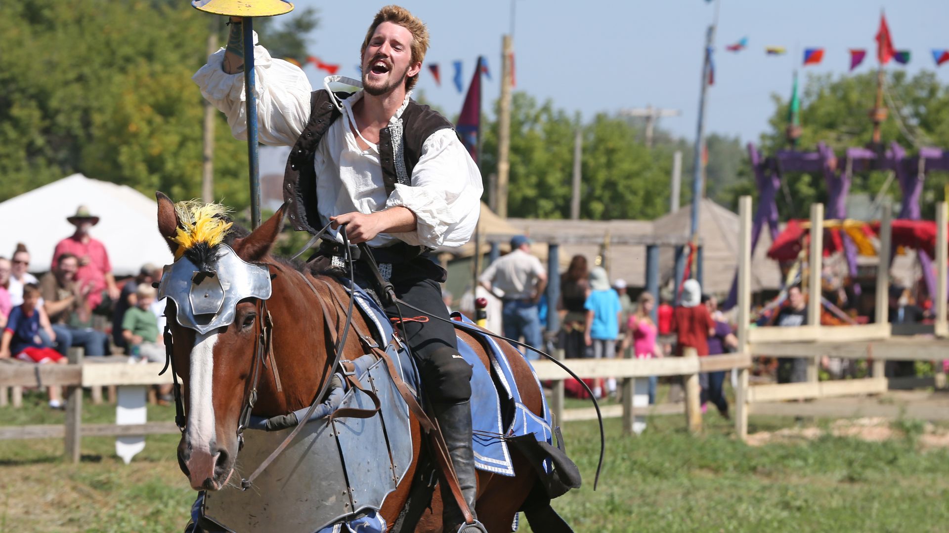A man dressed in Renaissance-era clothes rides a horse wearing armor through a field surrounded by spectators. 