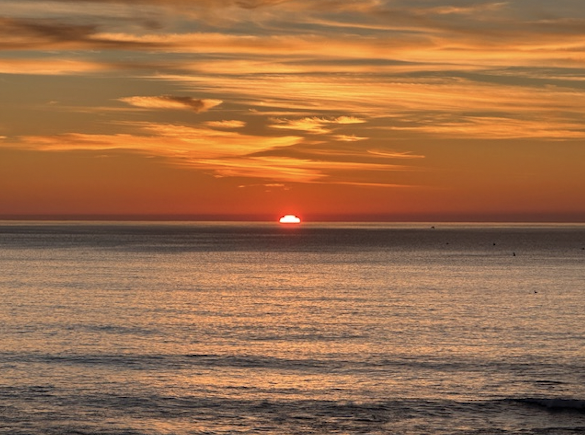 Sun setting over calm ocean with orange, yellow, and red hues in the sky and reflected on the water's surface under streaky clouds.