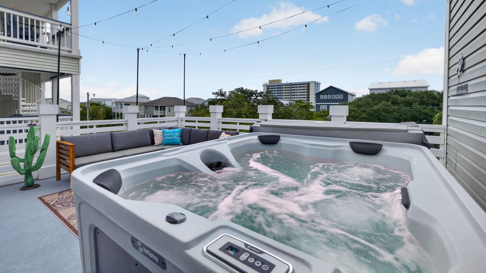 A rooftop deck with a large hot tub, gray sectional seating with blue cushions, string lights overhead, white railings, a cactus on the left, and distant houses under a blue sky.