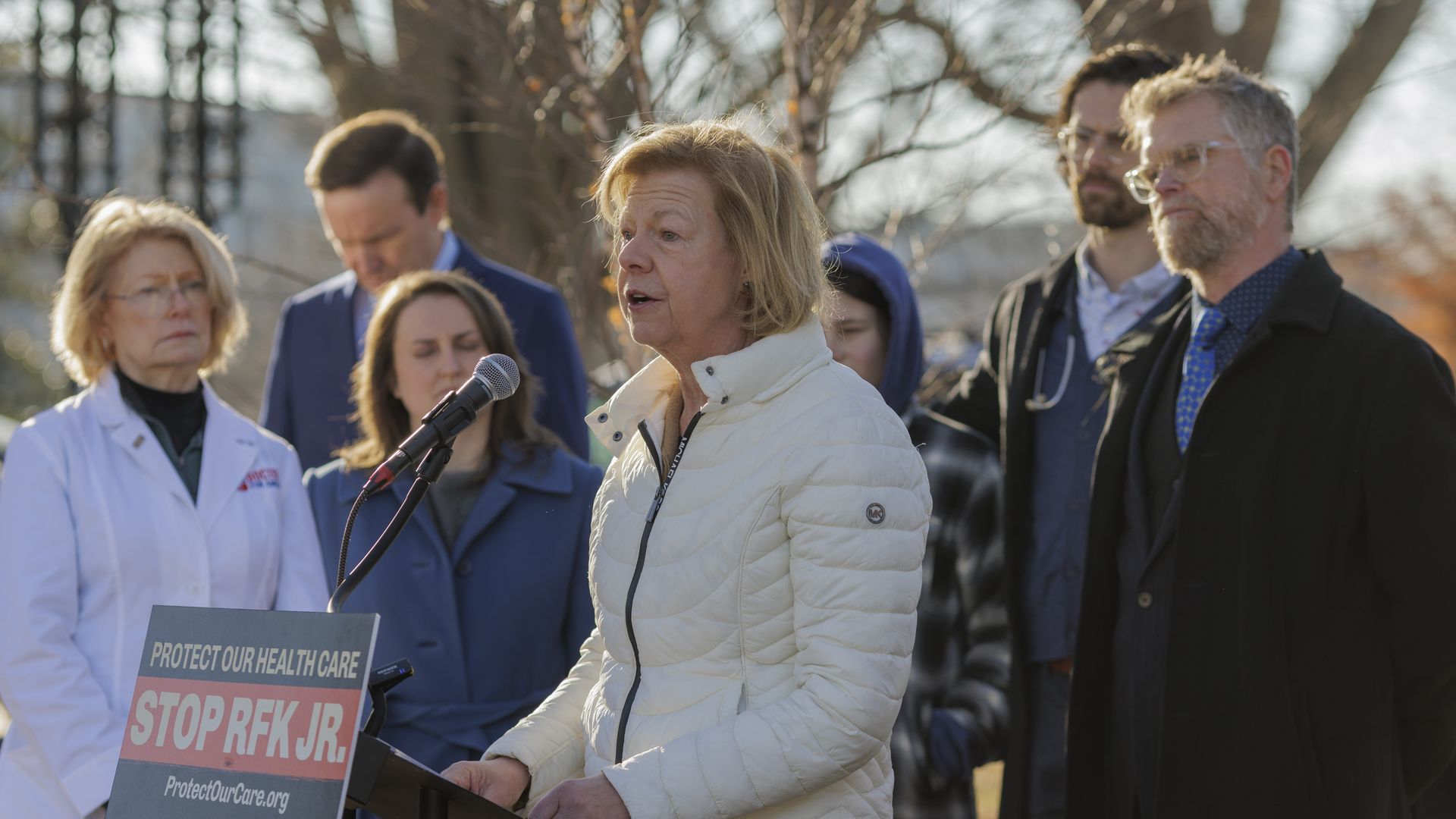 Tammy Baldwin speaks in front of a group of people from behind a lectern featuring a sign that reads "PROTECT OUR HEALTH CARE, STOP RFK JR."