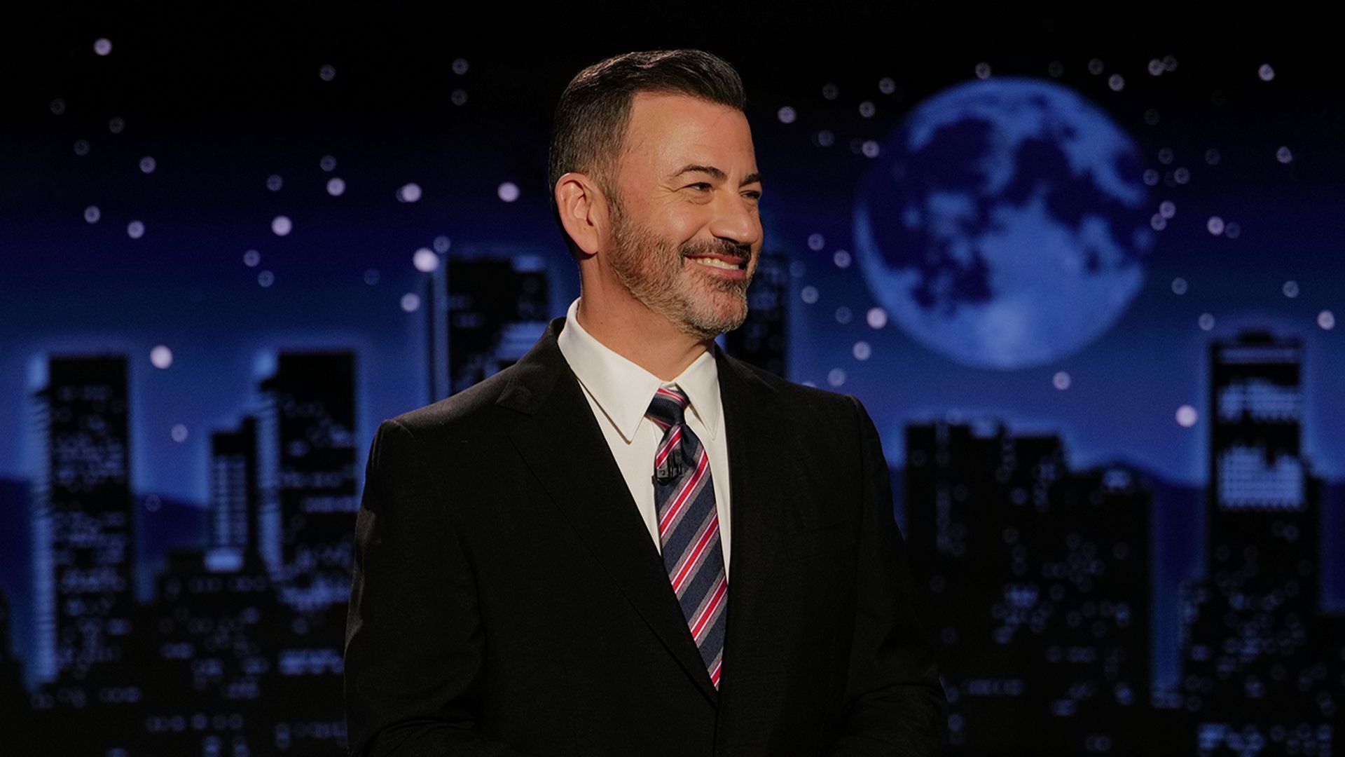 Jimmy Kimmel in a suit and tie smiles with his hands clasped while wearing a dark suit and a blue and red striped tie. Behind him is a night skyline background in blue tones.