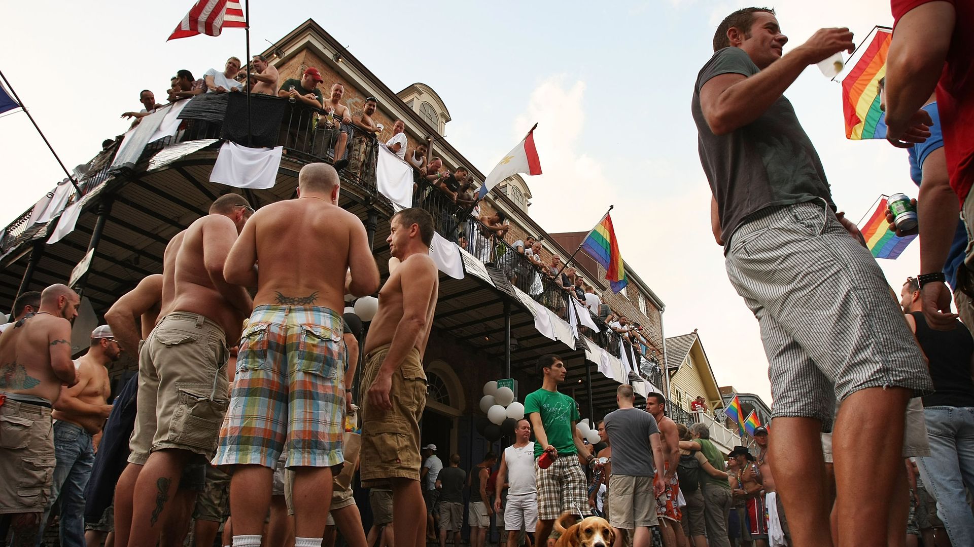 A French Quarter street is filled with revelers. Gay pride flags hang from a nearby balcony.