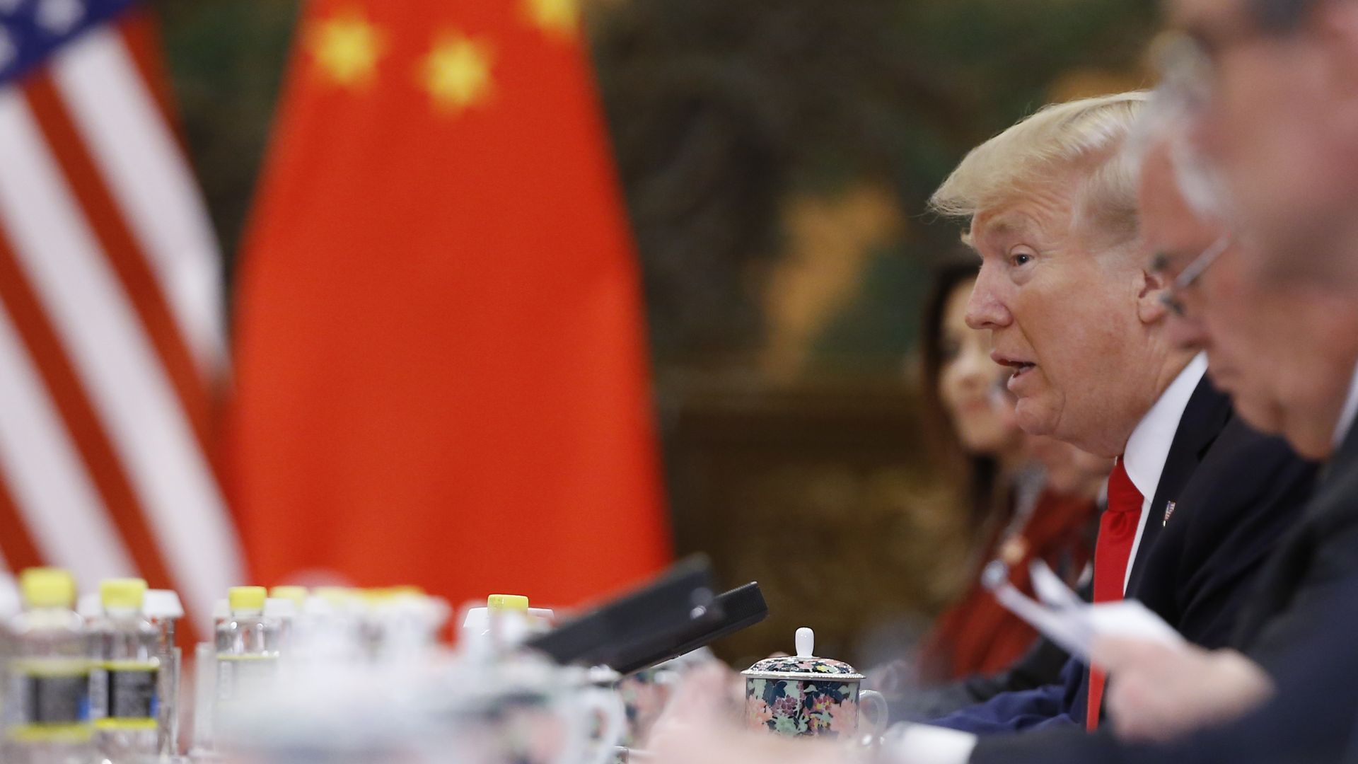 In this image, Trump sits at a table with other diplomats and political leaders. the Chinese flag is in the distance behind him.
