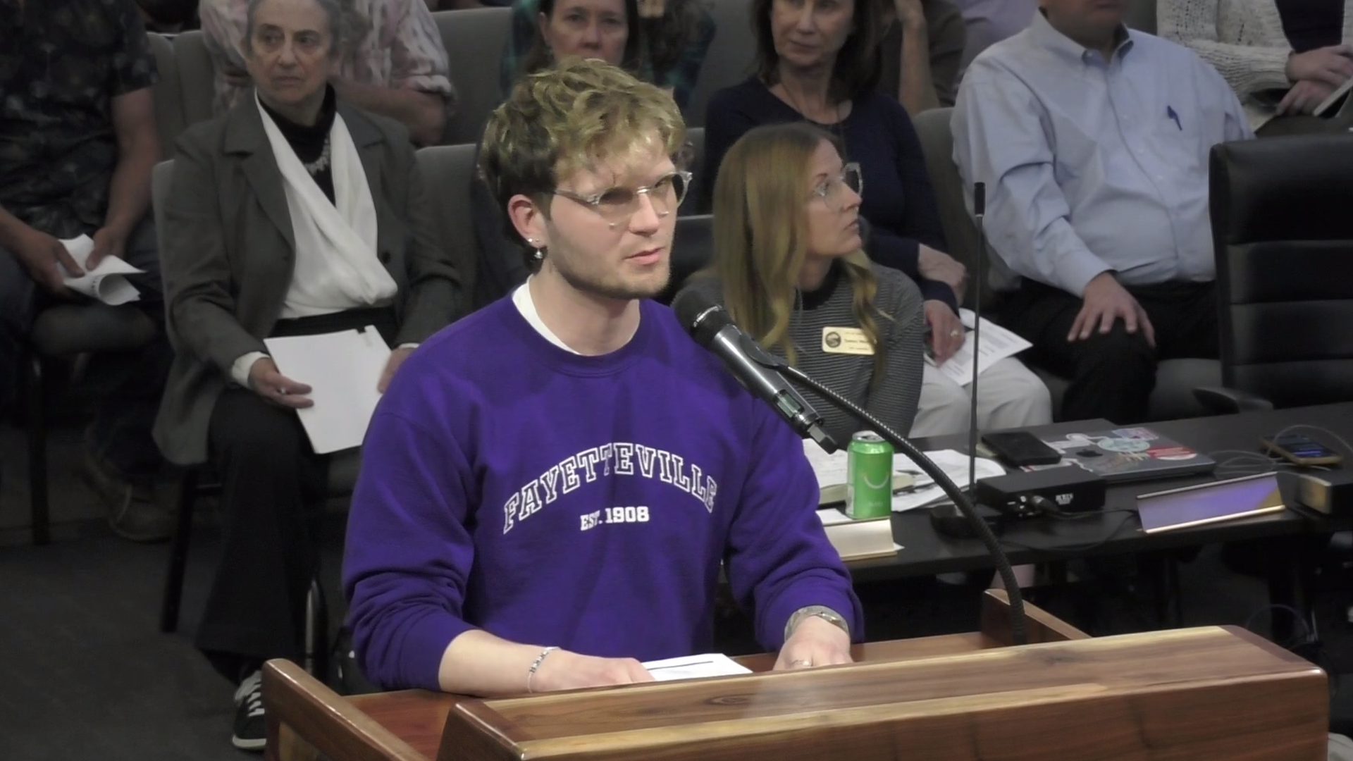 A young man in a purple "Fayetteville" sweatshirt speaks at a podium into a microphone, with an audience seated behind him.