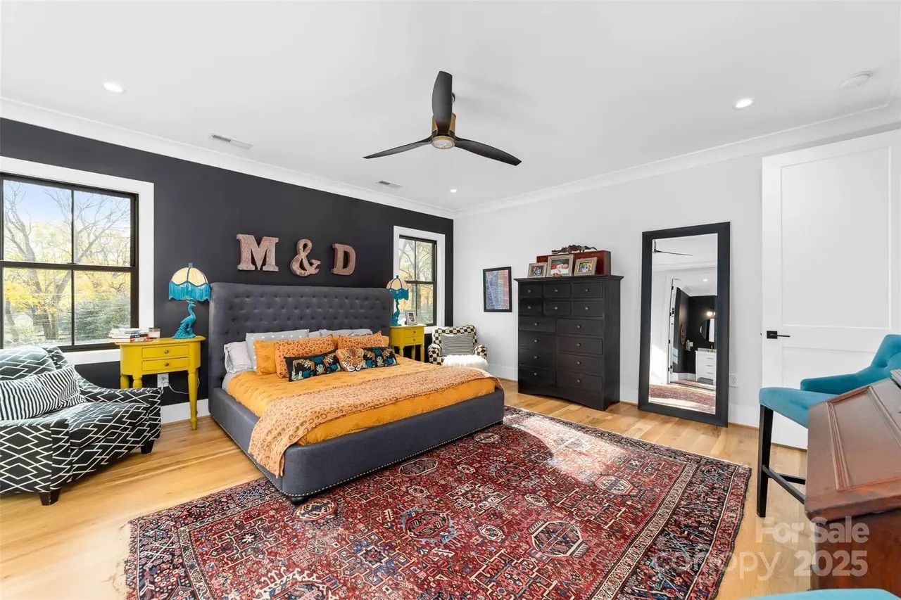Bright bedroom with a dark accent wall displaying large "M & D" letters, gray tufted bed with orange bedding, yellow nightstands with blue lamps, patterned armchairs, black dresser, full-length mirror, and large red rug.